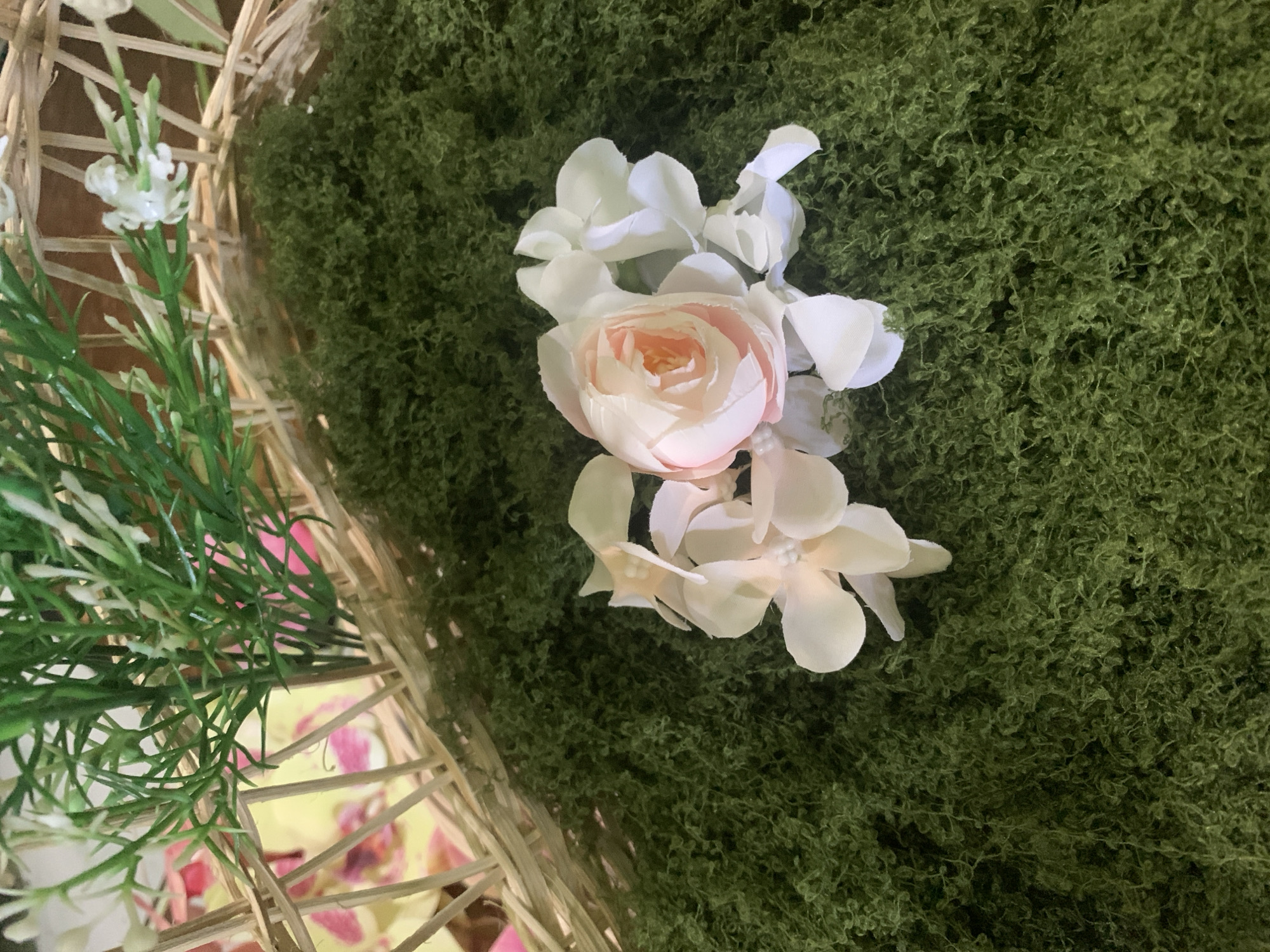 This image shows a close-up view of a decorative arrangement. In the foreground, a cluster of artificial flowers, featuring a pink and white rose and several white blossoms, rests on a bed of green artificial moss. In the background, a portion of a woven basket is visible, containing artificial greenery with small white flower buds. The overall scene appears to be staged as a decoration. There is no indication of a specific location in Kuala Pembuang, Indonesia, or any people present.