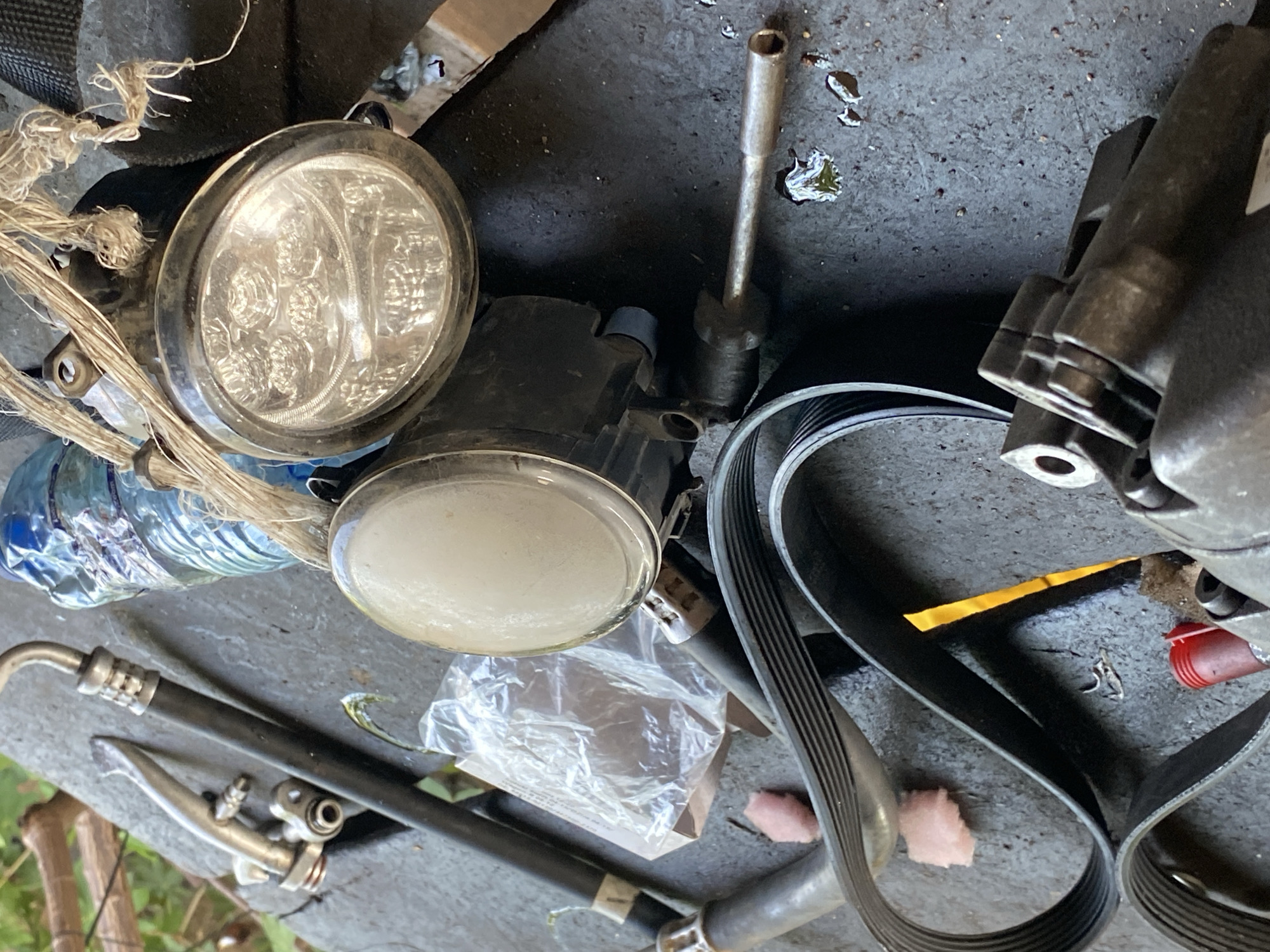 This image depicts a collection of automotive parts and other miscellaneous items scattered across a rough, gray surface, likely a workbench or the ground. The scene is set outdoors, as evidenced by the blurred green foliage visible in the background and the natural light.
Dominating the frame are several car parts. A prominent serpentine belt, a black rubber belt with multiple grooves, is coiled and draped across the surface. Nearby, a car's power steering pump or a similar component, made of dark gray metal, is partially visible on the left. Two circular light fixtures, possibly headlights or fog lights, are positioned towards the center and right of the image. The one on the right is a modern LED light, encased in a black housing and partially wrapped with coarse, light-brown twine. The other light fixture, to its left, appears to be an older halogen bulb type, with a clear glass lens.
Other objects in the scene include a clear plastic bottle containing a blue liquid, a length of black hose with metal fittings, a small red plastic cap, and two small, pink, foam-like pieces. There are also small metal shavings and some spilled liquid, possibly oil or water, on the gray surface.
The overall impression is one of disarray, suggesting that these parts are either awaiting installation, are being repaired, or have been discarded. The location is stated to be in the city of Jalingo, Nigeria.