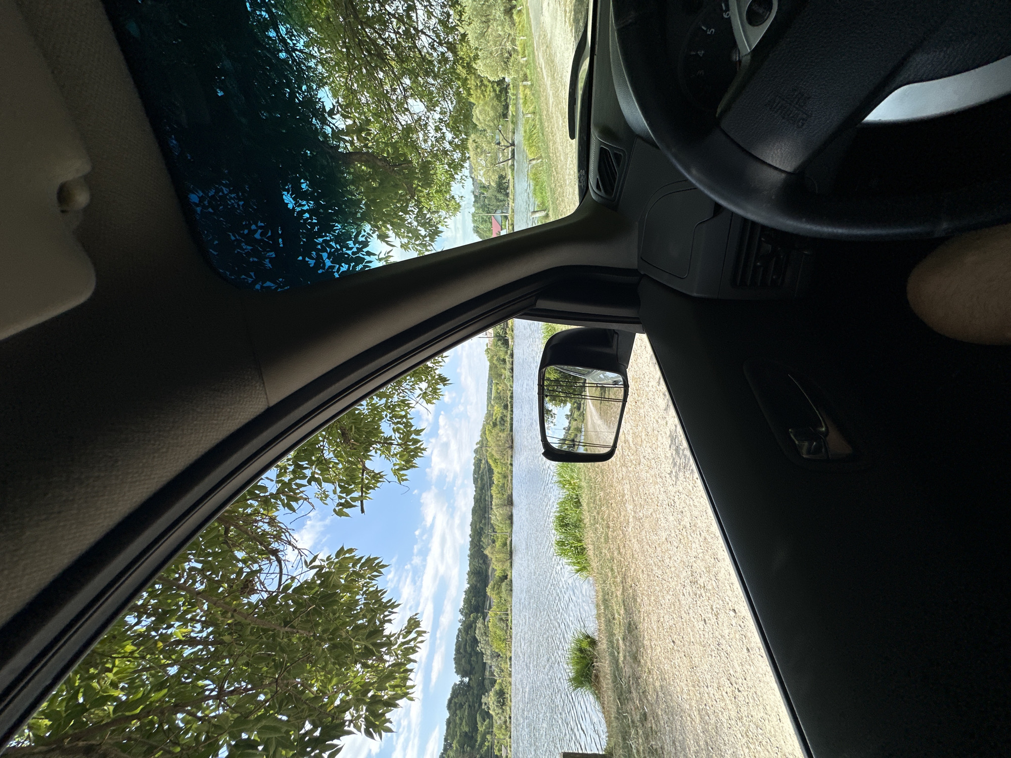 The image is taken from inside a vehicle looking out the driver's side window. The foreground shows the interior of the car, including a portion of the steering wheel, dashboard, and door panel. The window frames a view of a serene body of water, likely a lake or river, bordered by a grassy embankment and trees. The sky is blue with scattered white clouds. In the distance, a treeline is visible, with some buildings interspersed. A side-view mirror reflects a gravel path and more trees. The scene appears to be set in a natural environment, possibly a park or a rural area. The location in Makiv, Ukraine, is not discernible from the visual elements of the image alone.