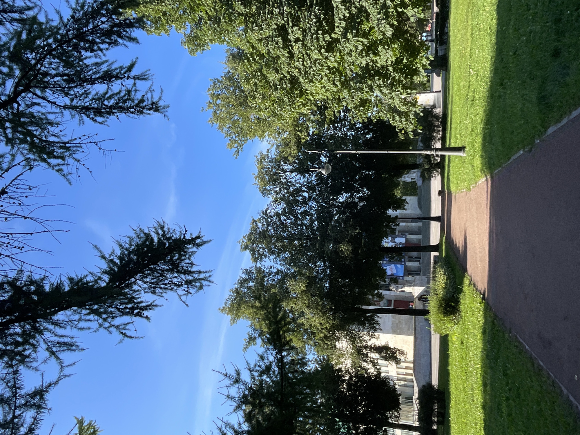 A pathway lined with green grass and trees leads toward a building in Saint Petersburg, Russia. The pathway is reddish-brown and curves gently to the right. On the left side of the pathway, there is a well-maintained lawn. On the right side, there are trees with lush green foliage and a lamppost. In the background, a large building with a light-colored facade is visible, along with a few people. The sky is a clear blue with a few wispy white clouds. The image is taken from a low angle, looking up towards the sky and the trees.