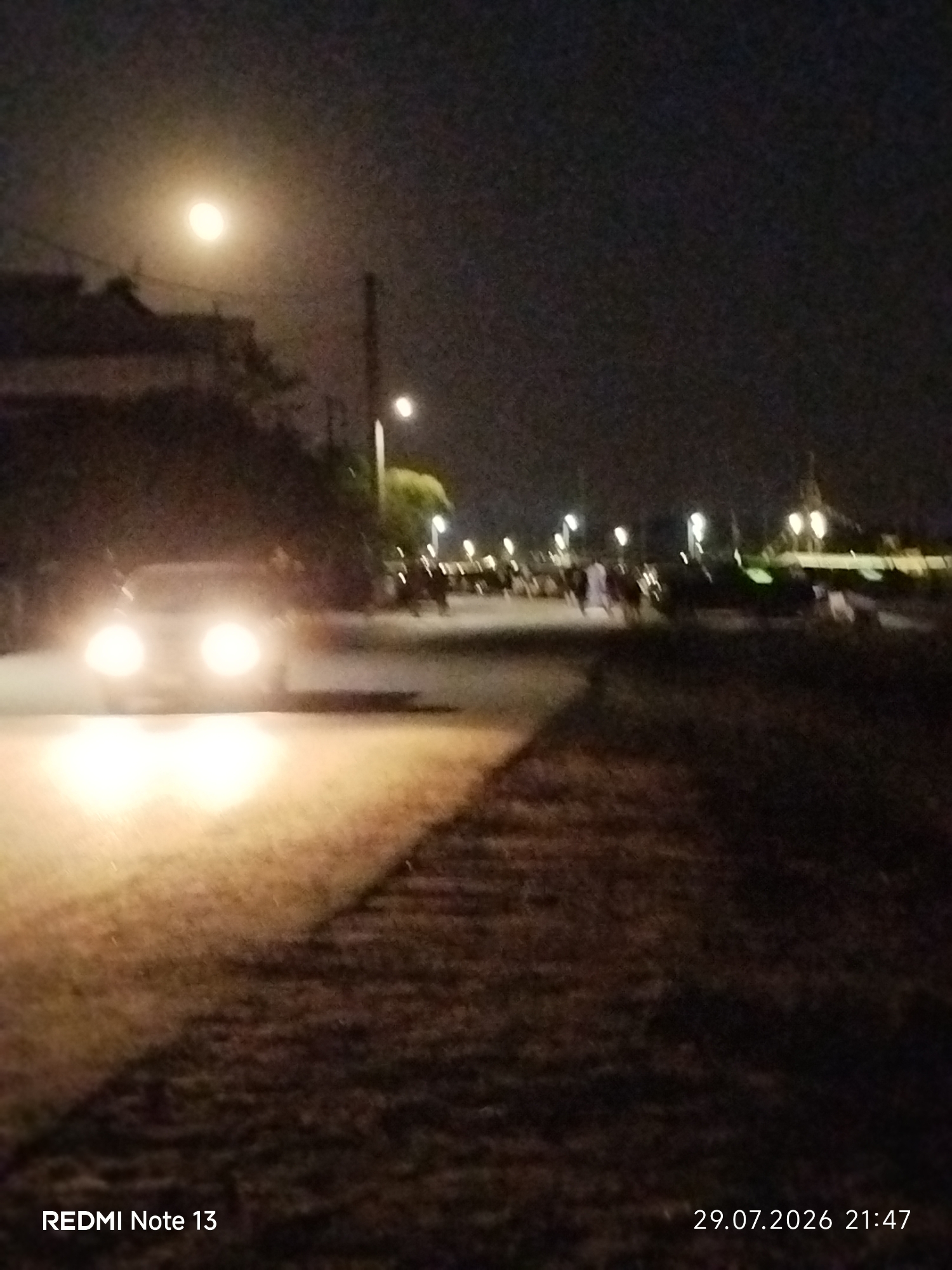 The image depicts a nighttime street scene, likely in the Sithonia Municipal Unit, Greece. A car with its headlights on is approaching, illuminating the road and the foreground. Several individuals, appearing as dark figures, are visible walking on the right side of the road. Streetlights dot the background, and a building structure is present on the left. The overall lighting is dim, with the car's headlights being the brightest element. The timestamp "29.07.2026 21:47" is present in the lower right corner, and "REDMI Note 13" is in the lower left, indicating the device used for capture.