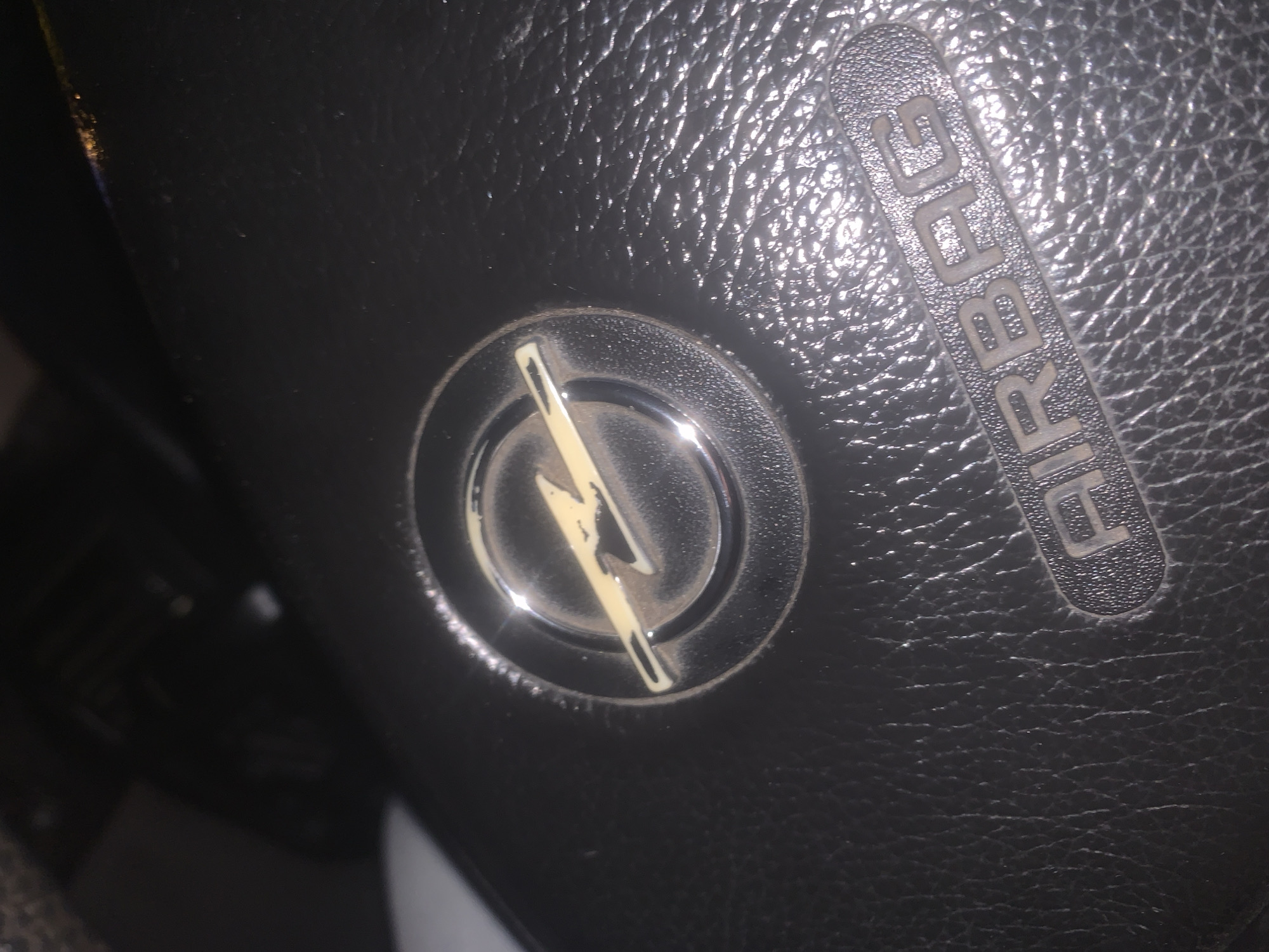 A close-up, high-angle interior shot depicts a vehicle's dark-textured steering wheel, located in the city of Gyumri, Armenia. Centrally positioned is a circular, metallic Opel emblem, featuring a light-colored lightning bolt. The emblem and the surrounding dark, textured material, consistent with a synthetic leather or plastic surface, exhibit visible accumulation of dust and minor surface wear. Below the emblem, the word "AIRBAG" is embossed in raised, block capital letters, also showing signs of dust and slight discoloration. Strong, localized reflections are present on the metallic emblem and the textured surface, indicating the presence of an immediate light source. The background is dark and out of focus, containing indistinct forms of other vehicle interior components.