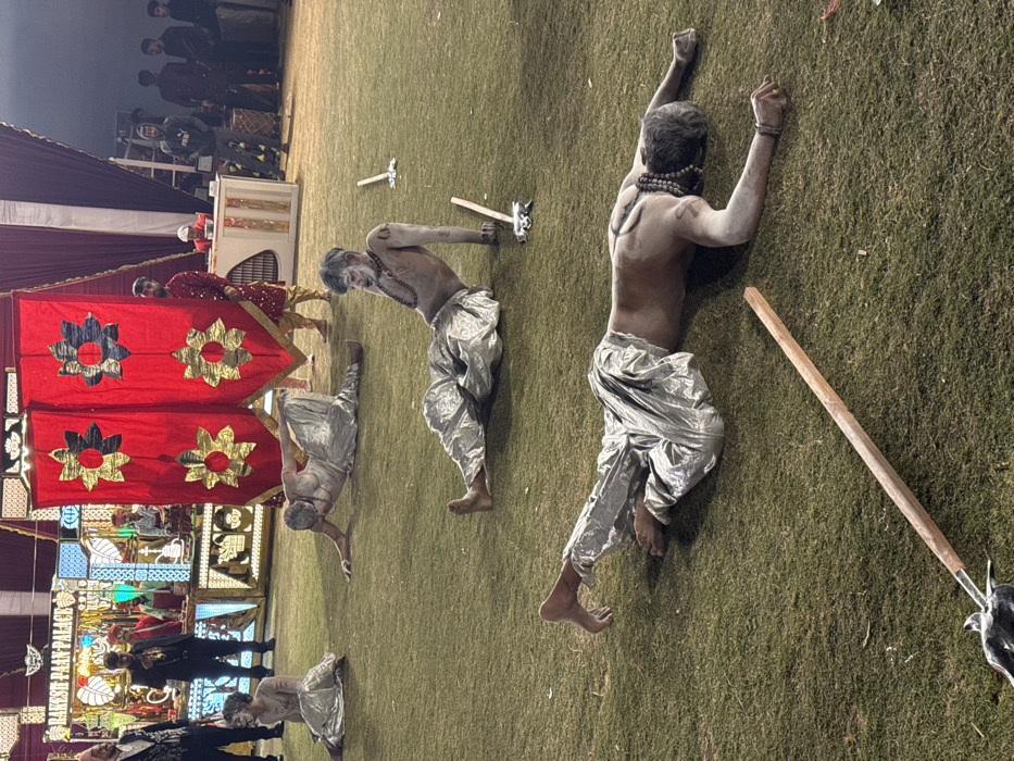 Four men in silver pants and white body paint perform on a grass surface, possibly during a cultural event. The men are in various yoga-like poses, with objects resembling a sword and a wooden stick near them. Red banners adorned with gold flower designs are positioned to the left. A group of people are seen in the background. The setting appears to be outdoors, potentially a stage or performance area.