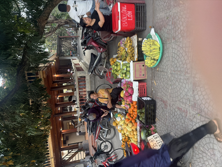 The image depicts an outdoor street market scene in Nam Hoa Lư Ward, Vietnam, likely during the late afternoon or early evening.
In the foreground, the lower body of a person wearing a dark blue long-sleeved shirt, dark pants, and sandals walks left, partially obscuring a red motorbike with license plate "35-H1 037.97".
The mid-ground features a vibrant fruit stall setup on patterned grey and brown interlocking pavers. Three women are actively present. To the left, an older woman with short grey hair, wearing a dark purple long-sleeved shirt and dark pants, is bent over a small silver digital scale, appearing to weigh produce. Next to her, a woman with short dark hair, wearing a yellow t-shirt and dark pants, sits on a low red plastic stool, looking towards the left foreground.
The fruit display includes large piles of yellow mangoes and orange tangerines/oranges directly on the ground. A small watermelon is also visible. In front of the woman in yellow, white styrofoam boxes hold pink dragon fruits with green tips. Black plastic crates contain green, unripe fruits, and various other green and yellow fruits are stacked on small platforms. To the right, another woman with short dark hair, wearing a black t-shirt, sits on a low stool, actively engaging in a transaction or preparing items, smiling towards the right. Next to her is a red cooler with white text "BÁNH LÒNG LỢN". Bunches of ripe yellow bananas are arranged on a small elevated surface, and a green plastic basket in the immediate foreground contains lighter yellow bananas.
Behind the woman in black, a man wearing a white long-sleeved shirt with a black logo and a black baseball cap stands, looking down towards her, suggesting an interaction.
Multiple motorbikes and one red bicycle are parked behind the fruit stalls. These include black, red, and silver models.
The background is dominated by a multi-story building constructed with traditional Vietnamese architectural elements, featuring a prominent wooden upper level with numerous dark wooden pillars, ornate balustrades, and a deep overhang. A wooden staircase is visible on the left side of this building. The ground level appears to be a concrete or stucco structure with an open porch where more motorbikes are parked. Large trees with green foliage are present, casting natural shadows.