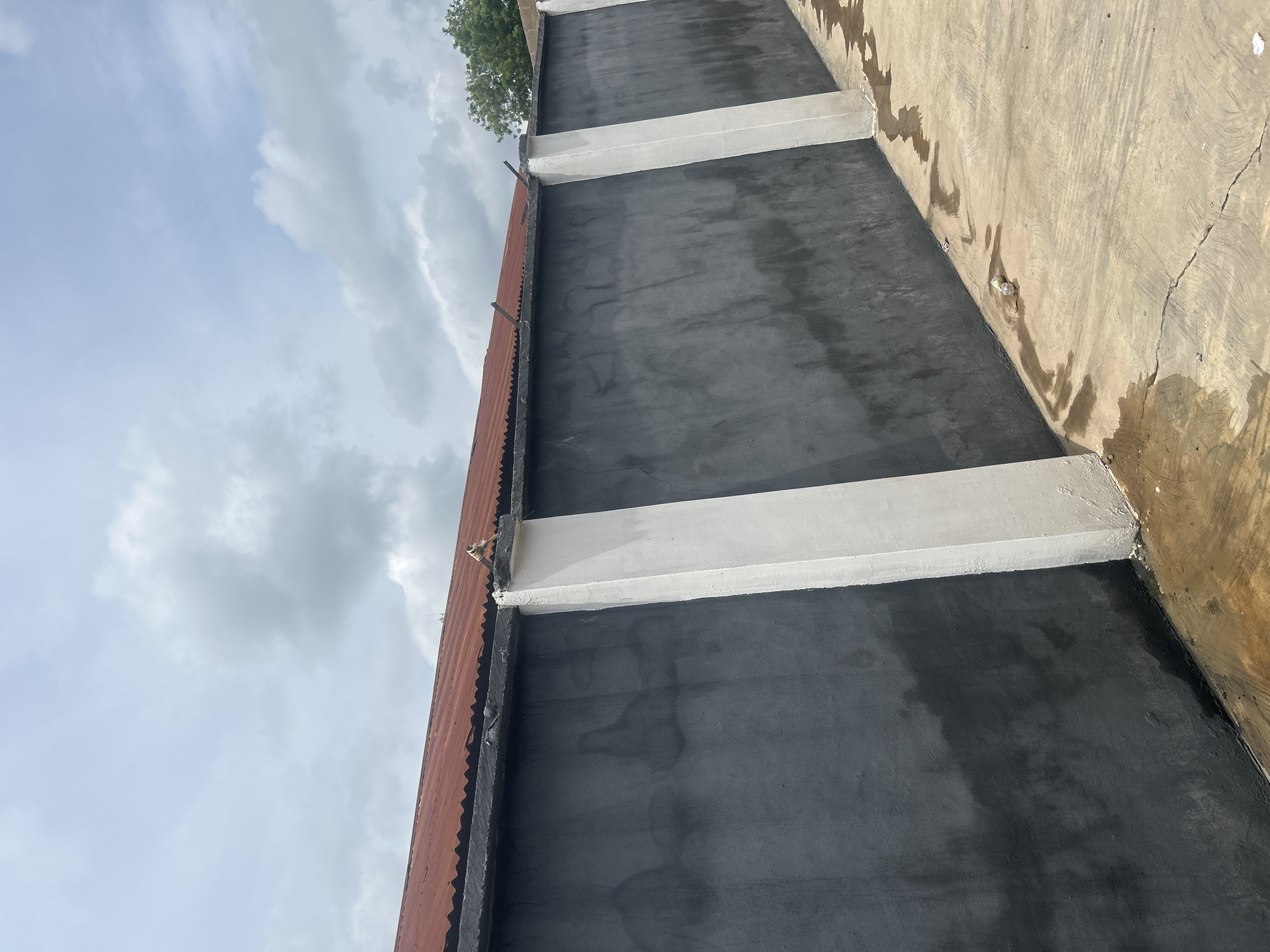 The image captures an outdoor scene in Ogbomosho, Nigeria, focusing on a dark gray, textured concrete wall that stretches diagonally from the lower left to the upper right. Two prominent, vertical white rectangular pillars are integrated into the wall, positioned at intervals along its length. The wall is topped by a corrugated, reddish-brown metal roof. Above the wall, the sky is partly cloudy, with patches of blue visible through the diffused light. The ground in front of the wall is a light brown, textured concrete surface with visible cracks and water stains, suggesting recent rainfall or cleaning. Scattered on the ground are a few pieces of litter, including what appears to be a crushed plastic bottle and some small paper fragments. The overall impression is of an exterior wall and surrounding area, possibly a boundary or part of a building's enclosure.