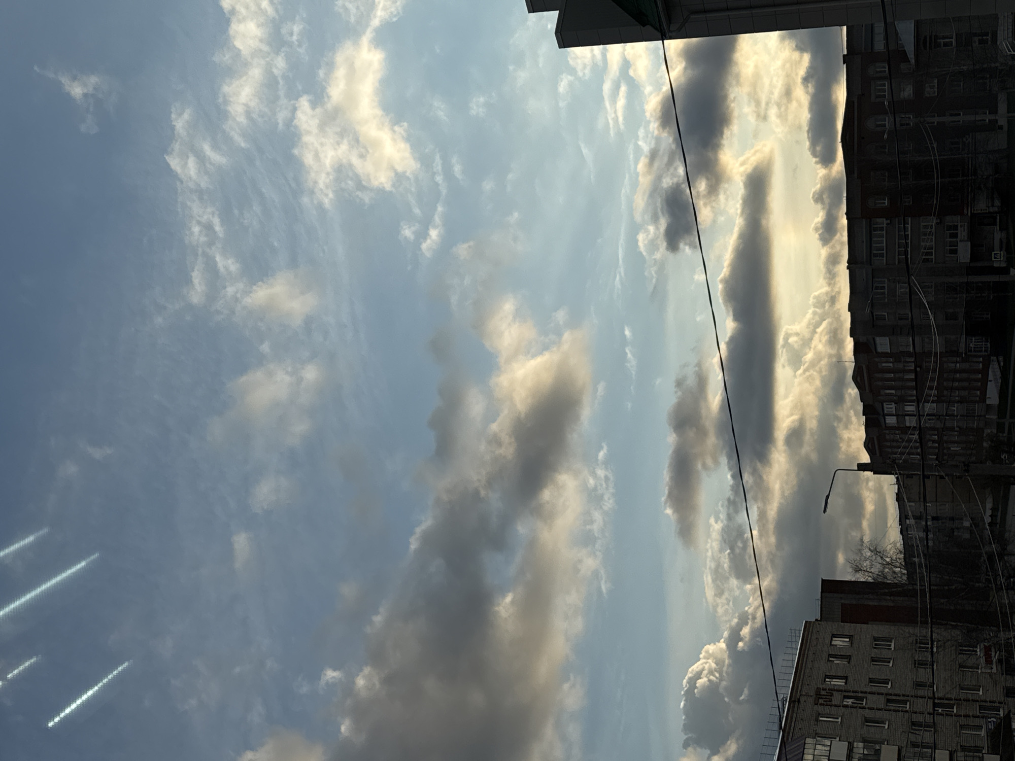 The image, captured in Chernushka, Russia, depicts a dramatic urban sky scene with buildings silhouetted against sunlit clouds. The sky dominates the frame, displaying a dynamic interplay of light and shadow across various cloud formations. Patches of pale blue sky are visible, particularly on the left, but much of the sky is filled with clouds ranging from wispy white cirrus-like streaks to dense, voluminous grey and dark clouds.
The sun, positioned low and behind some of the heavier clouds on the right, casts brilliant golden-white light on their edges, creating a striking backlighting effect. This illumination suggests either late afternoon or early evening. The varied cloud structures and lighting create a sense of depth and atmosphere.
On the right side of the frame, several multi-story urban buildings rise, appearing dark and largely in shadow against the brighter sky. Details like windows and architectural lines are discernible on their facades. A lamppost is visible, extending from behind one of the buildings. Numerous dark electrical wires and cables crisscross the upper right portion of the sky, characteristic of an urban environment. Bare tree branches can be seen behind one of the lower buildings, hinting at a colder season. On the far left, three distinct, bright diagonal streaks appear in the sky, likely reflections or lens flares.
The scene captures a moment of atmospheric beauty in a city landscape, emphasizing the natural elements of sky and light contrasting with man-made structures. There are no people visible, nor any discernible text.
