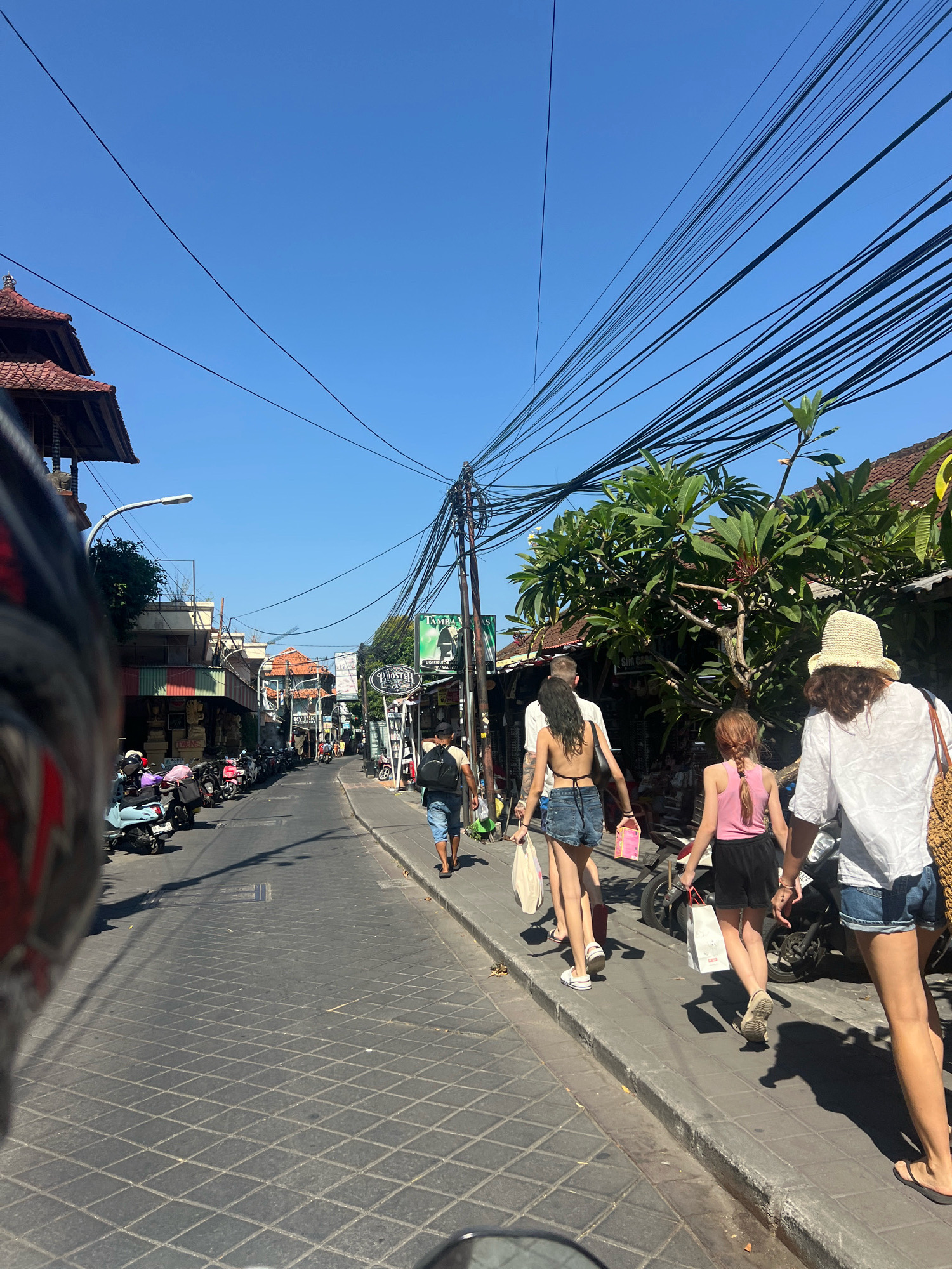 In Kuta, Indonesia, a street scene unfolds under a clear blue sky with numerous black wires overhead. The road is paved with patterned dark gray cobblestones and lined with shops on both sides. On the left, a row of parked scooters is visible. On the right side of the street, four individuals are walking away from the camera. A man in a light t-shirt and denim shorts with a black backpack walks ahead of a woman in a black bikini top and denim shorts. Next to her, a girl in a pink tank top and black shorts walks, followed by a woman wearing a white shirt, denim shorts, and a straw hat, carrying a woven bag. Multiple shops are open, with some displaying their wares. Trees and architectural elements, including tiled roofs, suggest a tropical urban environment.