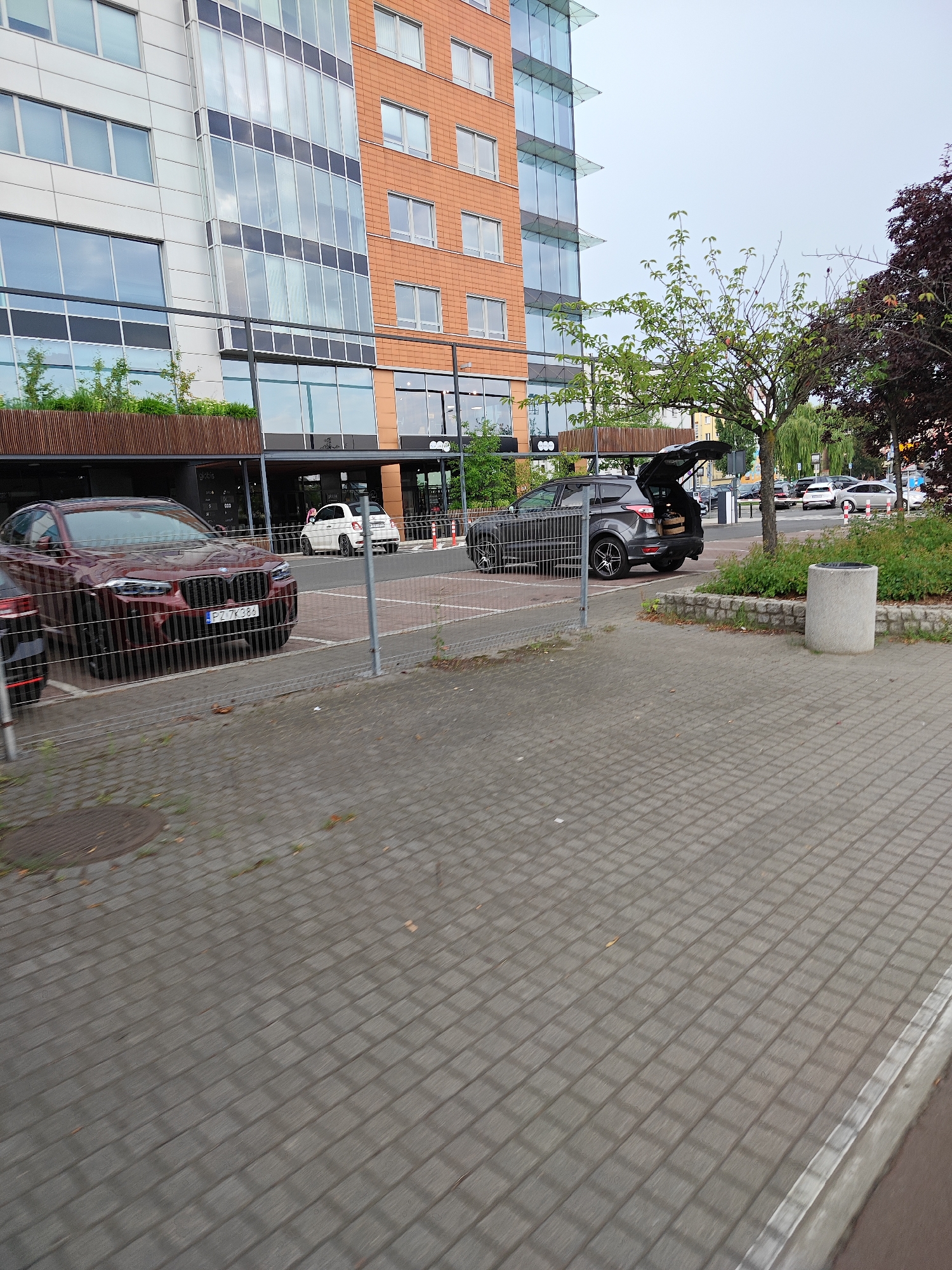 The image depicts a street-level view in the city of Poznan, Poland. In the foreground, a gray paved walkway with a diamond pattern extends towards the right edge of the frame. To the left, a metal fence separates the walkway from a parking area. Within the parking area, a dark red BMW SUV is parked, its front end visible and license plate reading "PZ 7K384". Behind the BMW, a white Fiat 500 is also parked. Further back, a dark gray SUV with its tailgate open is parked, containing some objects. The background features a modern building with a combination of white panels and orange brick facade, interspersed with large glass windows. Trees are visible on the right side of the image, including a larger tree with dark red leaves and a smaller green-leafed tree. The sky is overcast. The scene appears to be a commercial or residential area with parking facilities.