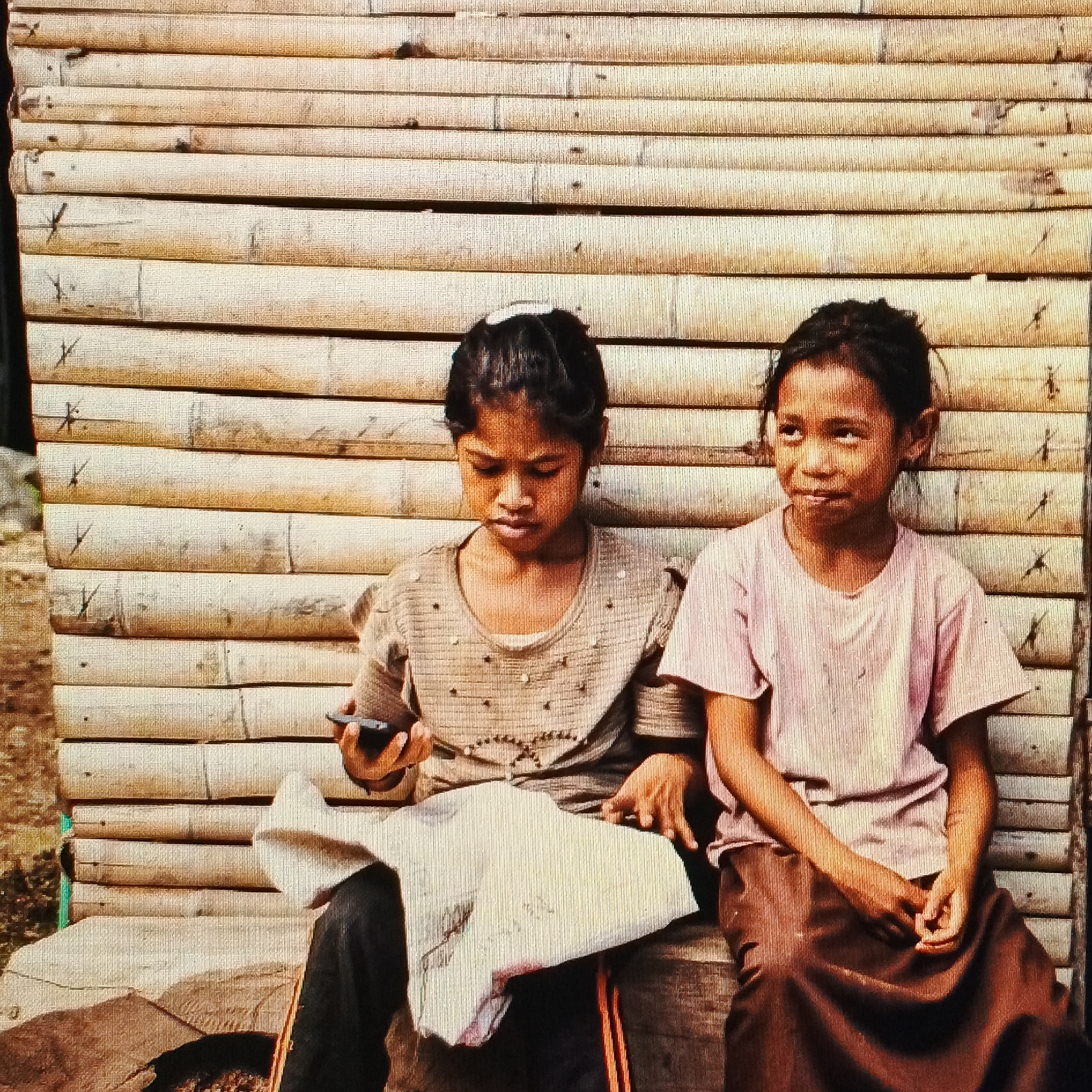 The image shows two young girls sitting on a wooden bench against a backdrop of horizontal bamboo poles. The girl on the left is wearing a light brown shirt with scattered embellishments and dark pants, and she is looking down at a smartphone in her hands. The girl on the right is wearing a light pink t-shirt and a brown skirt, and she is looking upwards and to her right with a slight smile. The environment appears to be outdoors, with the bamboo structure suggesting a rural or simple building. The lighting indicates it is daytime. The overall impression is a candid moment of childhood interaction with technology. There is no indication of the location being the city of Ende, Indonesia.