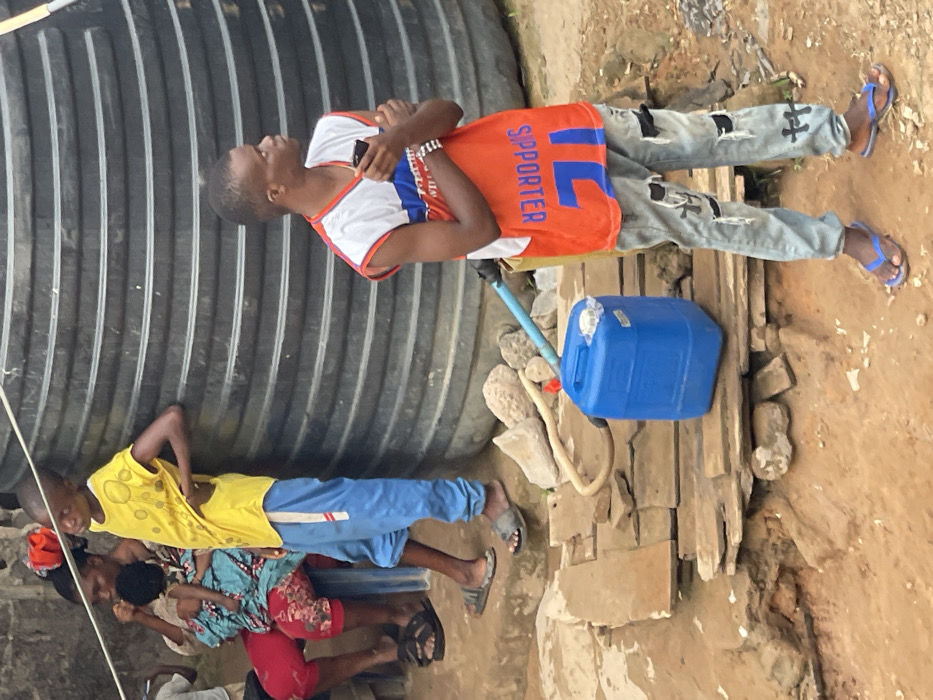 Two people stand near a large corrugated metal tank, one wearing a colorful shirt with "SUPPORTER" on the back and ripped jeans, the other in a yellow shirt and blue pants. A blue jerrycan sits on wooden planks with a hose leading to the tank. Below, a woman in a floral print shirt and red pants crouches, and a child in yellow and blue stands beside her. Another person is partially visible, wearing a white shirt. The ground around them appears to be a mix of dirt, wood, and rocks.