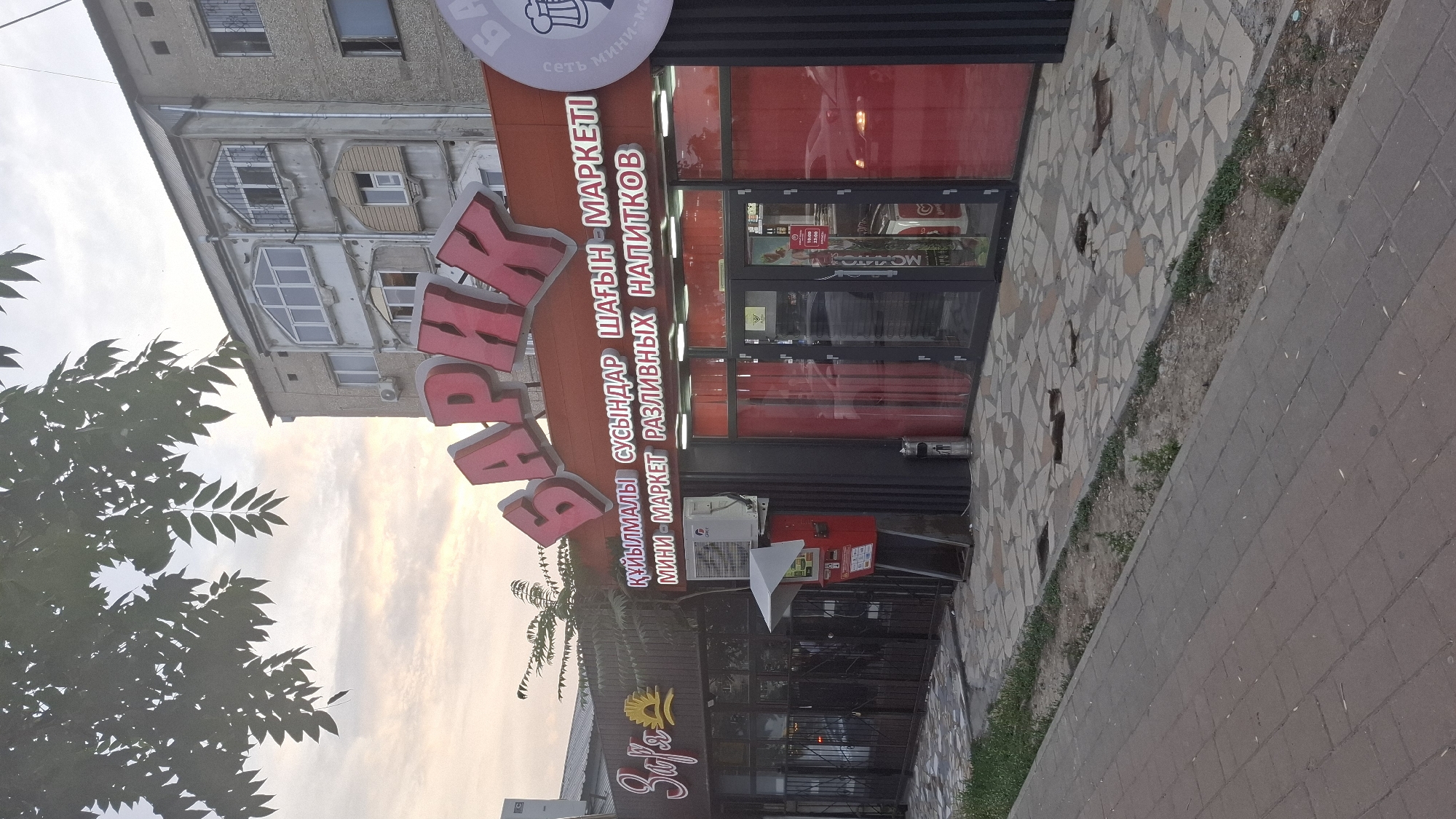 The image shows the exterior of a commercial establishment in Shymkent, Kazakhstan, with a prominent red and orange sign displaying the name "БАРИК" in large, stylized Cyrillic letters. Below this, smaller text in Cyrillic reads "ҚҰЙЫЛМАЛЫ СУСЫНДАР ШАҒЫН - МАРКЕТІ МИНИ - МАРКЕТ РАЗЛИВНЫХ НАПИТКОВ". To the left, another sign with the script "Zafr" and a stylized sun logo is visible, likely for an adjacent business.
The building appears to be a multi-story structure with architectural elements characteristic of Soviet-era construction, including a balcony with decorative metalwork and arched windows. The ground floor features a storefront with large glass doors and windows, revealing a bright red interior. An air conditioning unit is mounted on the exterior wall, and a red metal box on a stand, possibly a post box or information kiosk, is situated near the entrance. The sidewalk in front of the establishment is paved with grey bricks and irregular stone tiles, with patches of dirt and grass along the edge. The scene is illuminated by the soft light of what appears to be dawn or dusk, with a cloudy sky overhead and trees with green foliage visible in the upper left corner.
