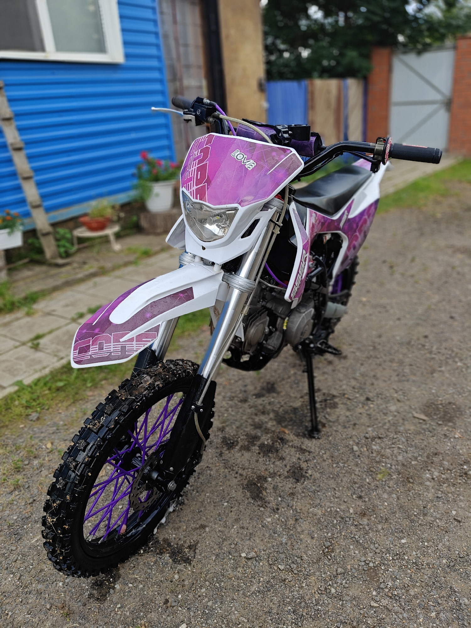 The image depicts a purple and white dirt bike parked on a gravel surface in an outdoor setting. The dirt bike is the primary subject, angled from the front right, showcasing its tires, suspension, and decorative graphics. The background features a blue corrugated building, a ladder leaning against it, potted plants, and a hint of greenery and fencing. The overall scene suggests a casual or recreational environment, possibly a yard or a rural area.