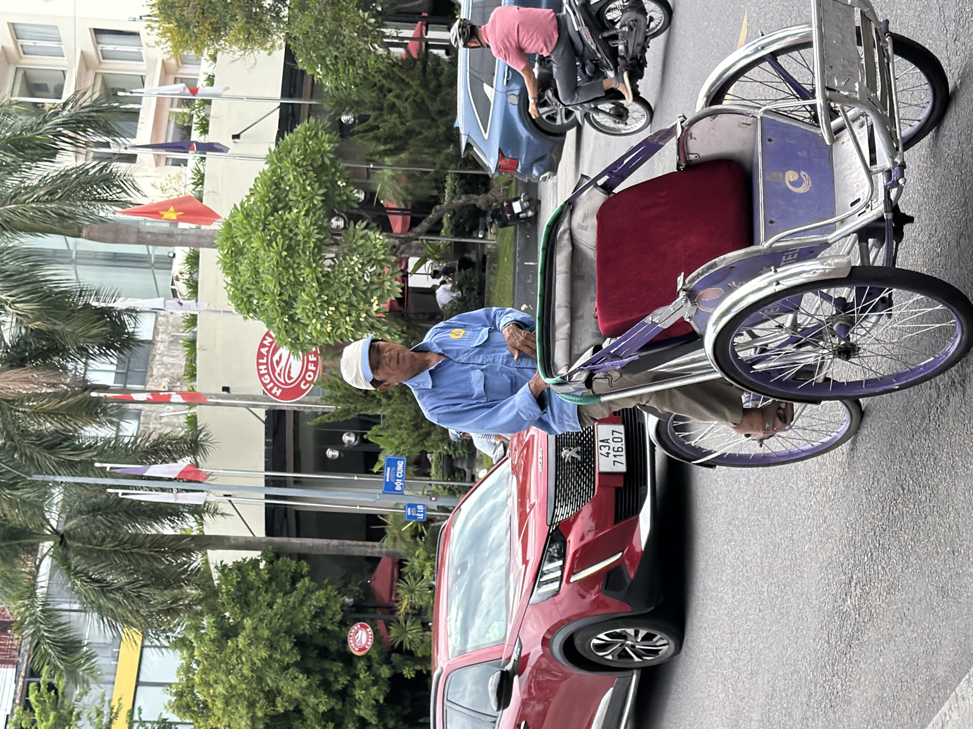 A man is pushing a purple and silver cyclopousse with a red seat on a street in Thuận Hóa Ward, Vietnam. He is wearing a light blue long-sleeved shirt, khaki pants, and a white cap. A red Peugeot SUV is parked behind him to the left, and a blue SUV is to the right. A person on a motorcycle is visible in the background. There are palm trees and buildings lining the street. Several flags are also visible. The sky is overcast.