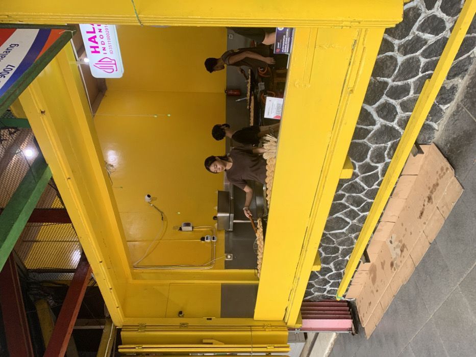 The image shows a food stall in Malang, Indonesia, located at coordinates -7.968, 112.628. The stall has a bright yellow exterior with a stone base. A woman is inside the stall, preparing food with a spatula. There are two other people in the background, one standing and another sitting. The stall has a sign that says "HALAL INDONESIA" with a phone number. The scene is likely a street food stall, offering various food items. The image is taken from a slightly elevated angle, showing the stall's exterior and the surrounding area.