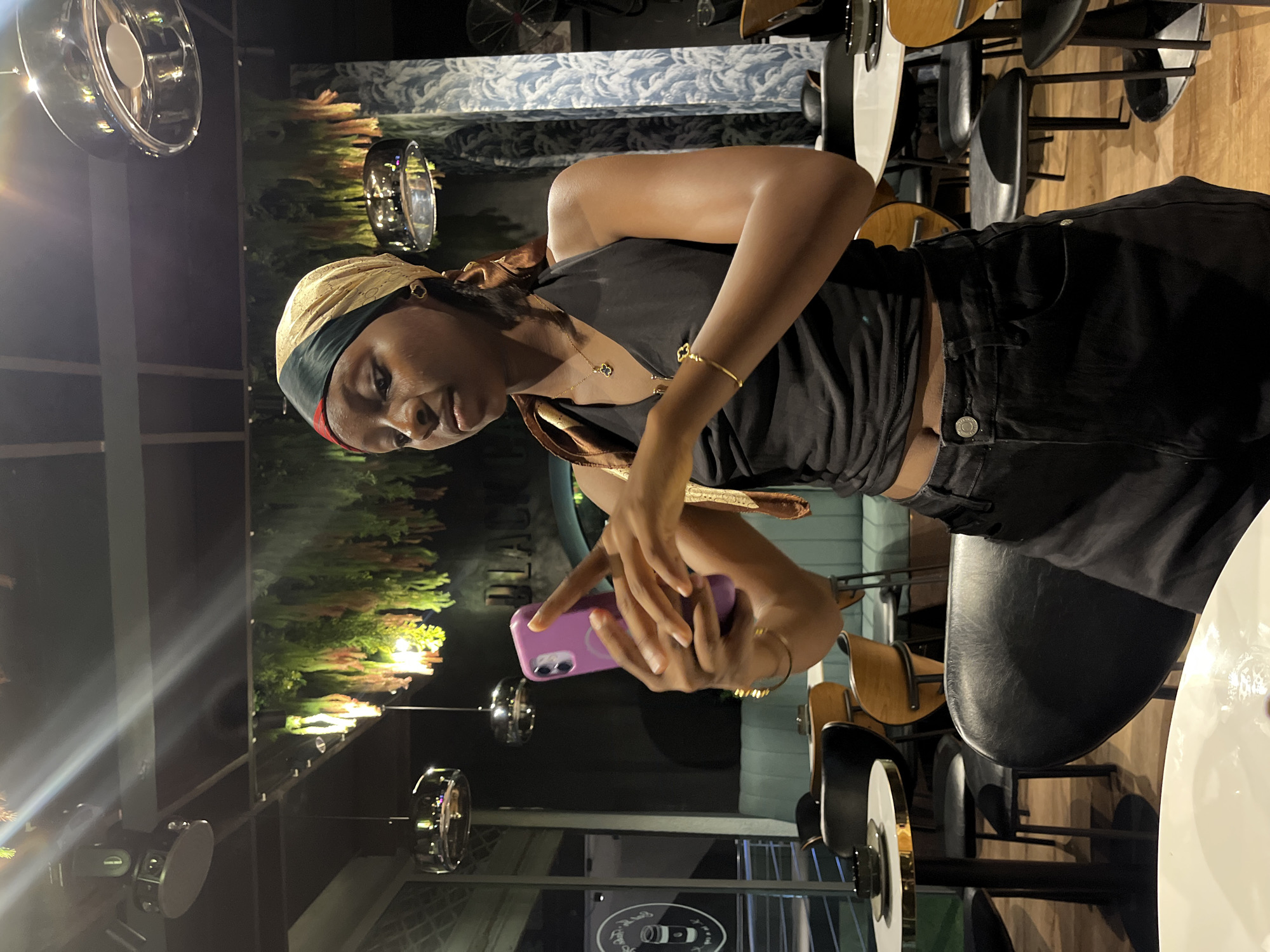 A young Black woman stands in a dimly lit restaurant, holding a purple smartphone in her hands and looking down at it. She is wearing a black halter top, black shorts, and a gold and green headscarf. She has gold jewelry on her wrist and neck. The restaurant has black chairs and tables, with some greenery hanging from the ceiling. The woman is positioned in the foreground, with the restaurant's interior visible in the background. The image appears to have been taken in the city of Ado, Nigeria.