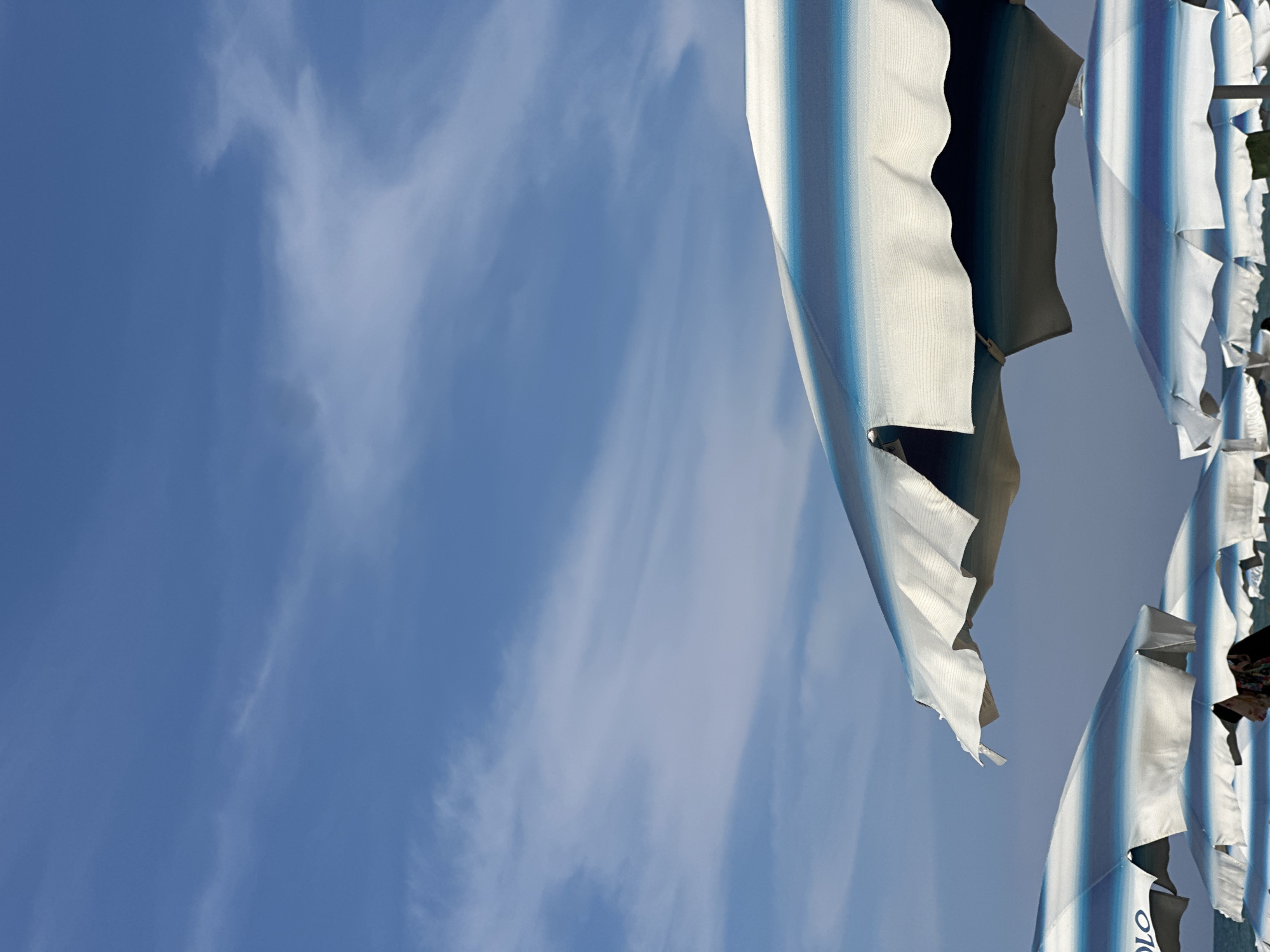 The image shows several beach umbrellas with blue and white stripes against a blue sky with wispy clouds. The umbrellas are partially furled and appear to be made of fabric. One umbrella on the right has text on it that appears to be "Polo". The image is taken from a low angle looking up at the sky and umbrellas. The lighting suggests it is daytime, likely sunny given the clear sky with some clouds. The scene appears to be a beach or a similar outdoor recreational area. No people are visible in the image.