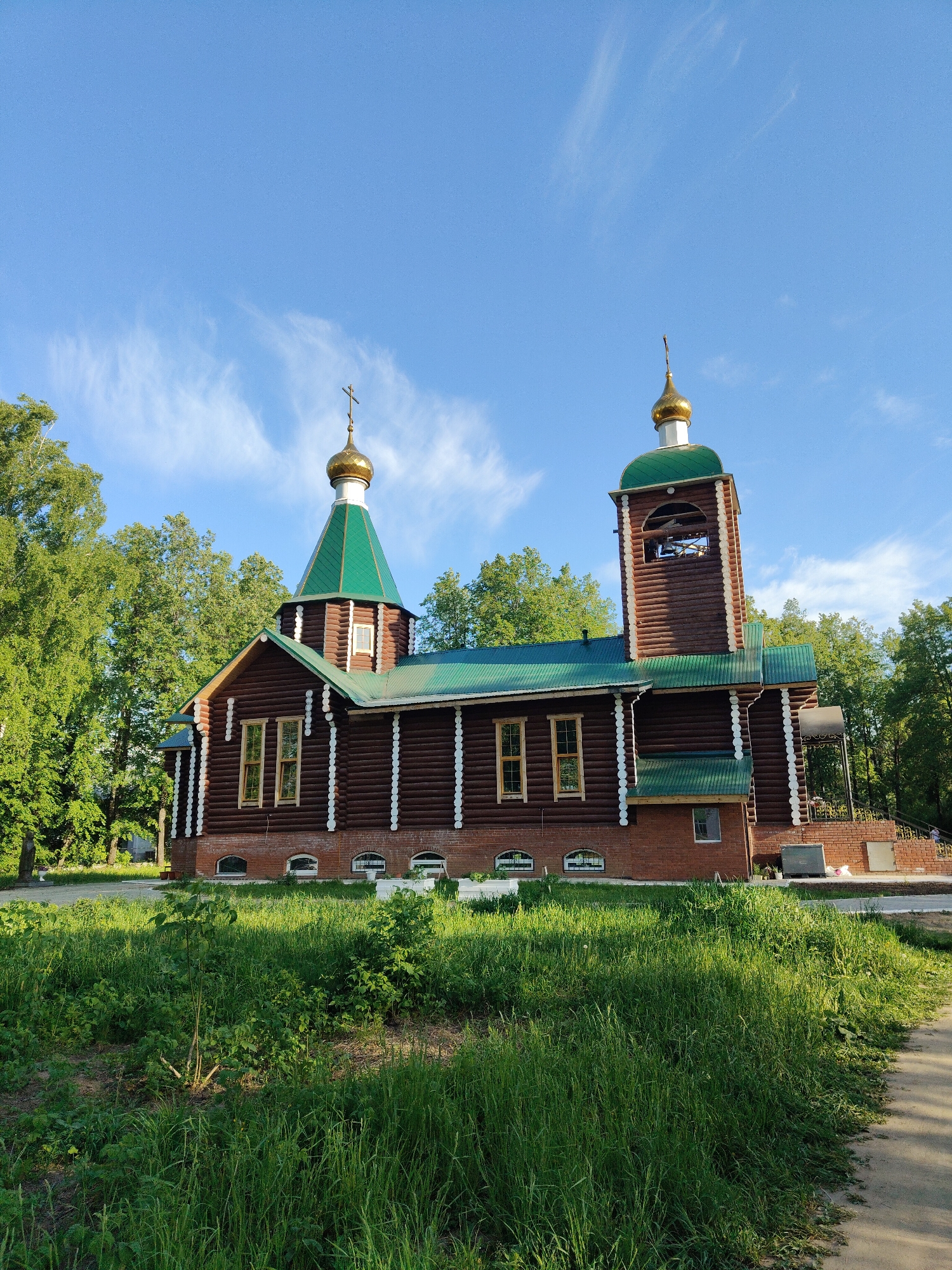A full shot shows a wooden church in Unknown, Unknown. The church is made of brown logs with white trim and a green roof. It has two towers, one with a gold dome and cross, and the other with a bell tower. The church is surrounded by green grass and trees. A sidewalk is on the right side of the image. The sky is blue with some wispy clouds.