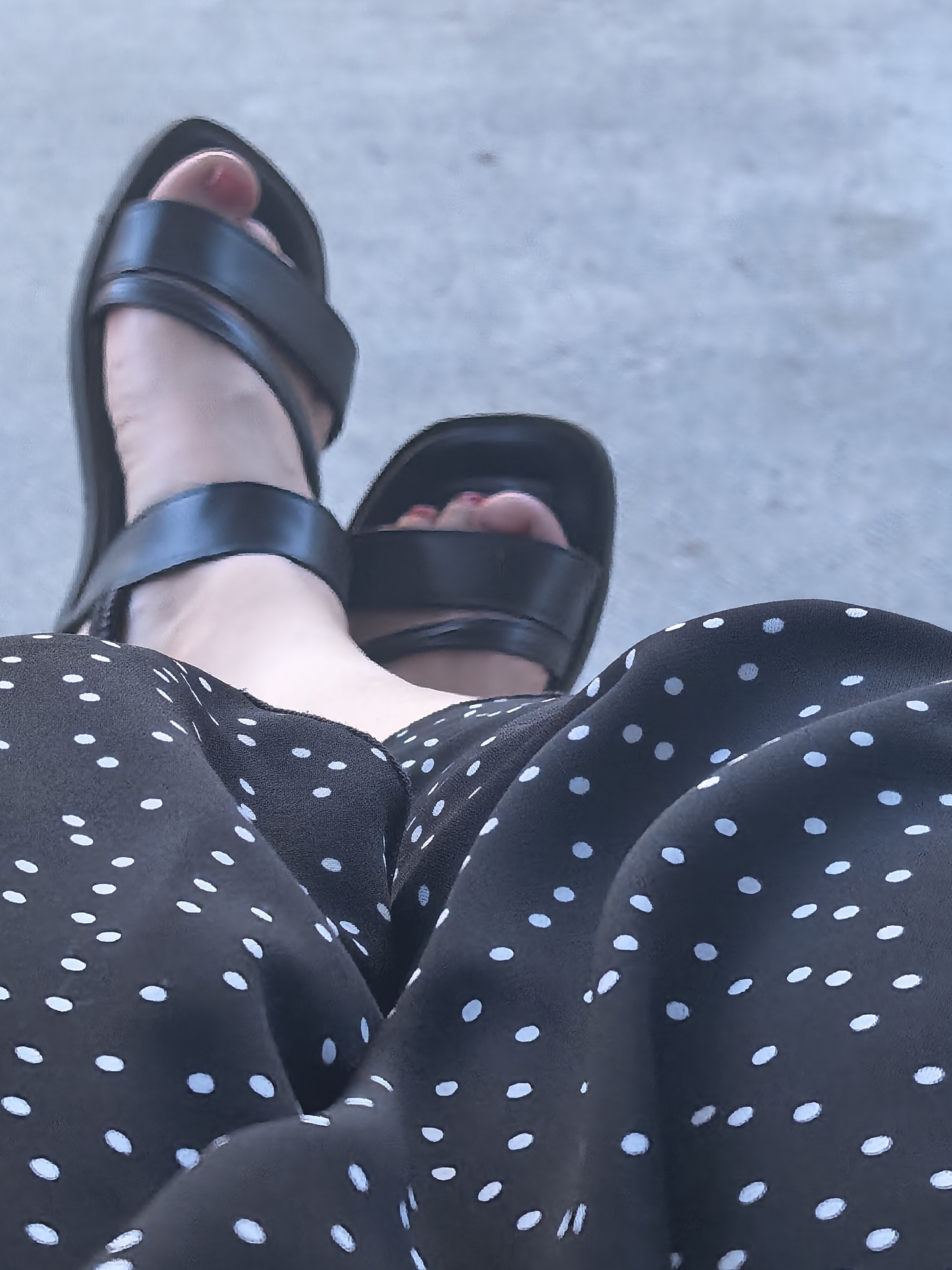 The image shows a close-up view of a person's feet wearing black sandals and a black and white polka dot dress. The sandals have thick straps that cross over the toes and around the ankle. The dress fabric is flowing and appears to be made of a light material. The polka dots are small and white, scattered across the black fabric.
The person's toenails are painted red, adding a pop of color to the otherwise monochromatic scene. The background is blurred and appears to be a light gray, possibly concrete or pavement. The lighting suggests it is daytime, but the specific time of day and weather are not discernible from the image.
The angle of the shot, looking down at the feet, implies that the person is either sitting or has their legs crossed. There is no visible text in the image. The overall impression is casual and relaxed. The focus on the feet and the dress suggests an interest in fashion or personal style. The image is cropped tightly, drawing attention to the textures and patterns of the clothing and footwear.