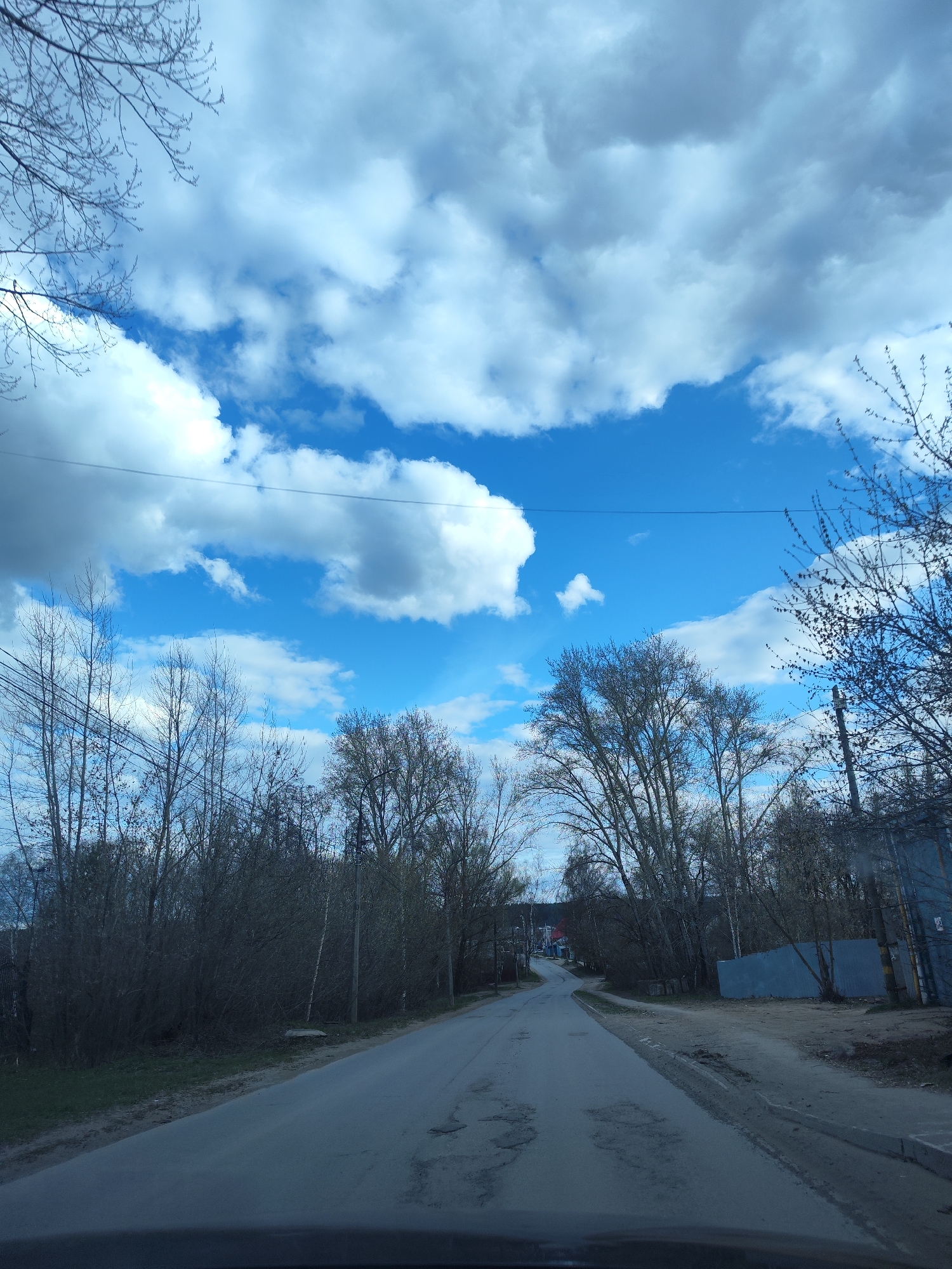 This image captures a daytime view from inside a vehicle traveling along a paved road in what appears to be a residential or semi-urban area of Chaikovsky, Russia. The road shows signs of wear, including several potholes, and curves gently into the distance.
Both sides of the road are lined with deciduous trees, most of which are bare or just beginning to bud, indicating either early spring or late winter. On the right side, beyond a dirt shoulder, a light blue or grey corrugated fence or wall is visible, behind which more trees stand. Power lines stretch across the upper part of the frame, traversing the clear blue sky.
The sky is a prominent feature, filled with large, bright white cumulus clouds, suggesting clear and pleasant weather. No people or specific activities are visible, contributing to a sense of quietude in the scene. The overall impression is of a journey through a transitional landscape under a bright, cloud-dotted sky.
