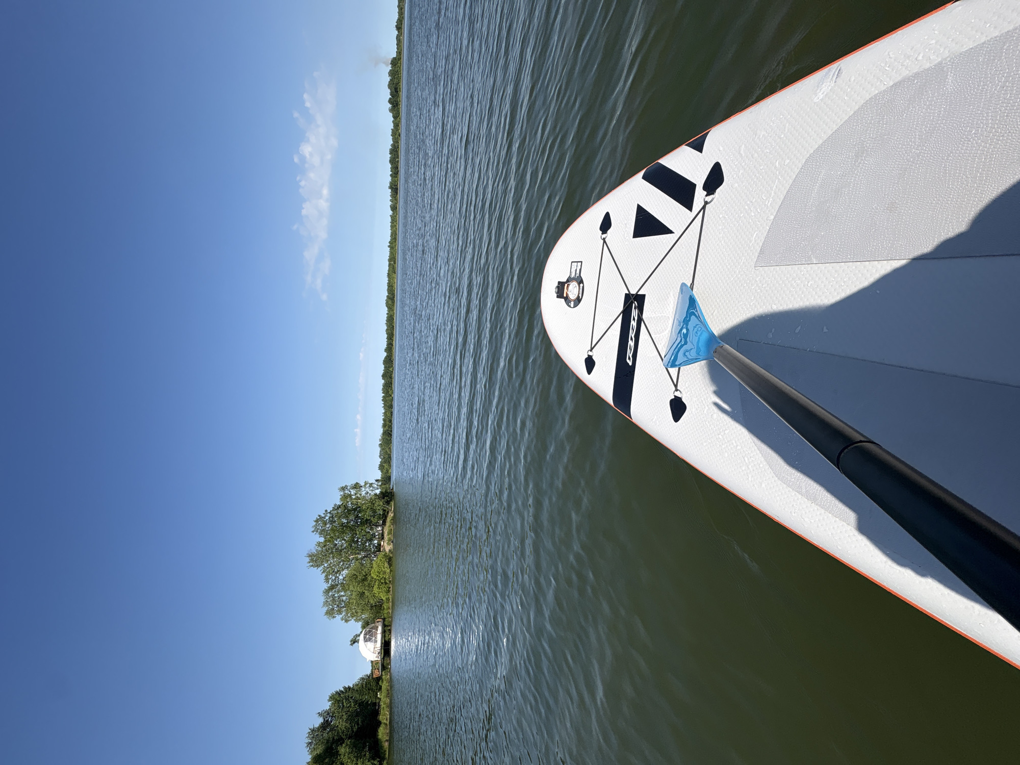 The image is a first-person perspective of someone on a paddleboard on a calm body of water, likely a lake or bay, under a clear blue sky. The paddleboard in the foreground is white with orange trim and black graphics, with a paddle partially visible. In the distance, across the water, is a tree-lined shore with a distinctive white, dome-shaped structure, possibly a geodesic dome home or glamping pod, situated on a small island or peninsula. The water is a murky green, with small ripples indicating gentle movement. The overall scene is peaceful and outdoorsy. The location is described as Razdolnoye, Russia, though this specific information cannot be verified solely from the image.