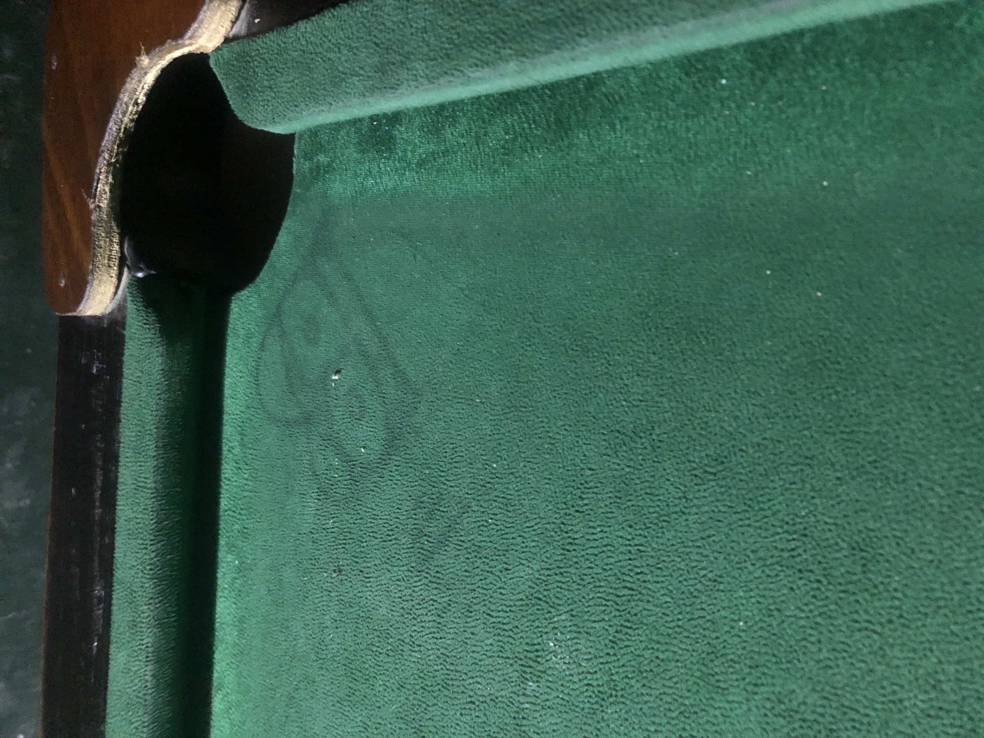 The image shows a close-up, oblique view of a green felt pool table surface, focusing on the area near a corner pocket and the edge of the table. The felt is textured with a ribbed pattern. A faint, dark circular or oval mark is visible on the felt, possibly from a pool ball or a stain. The wooden rail of the pool table is visible at the top and right edges, with the corner pocket area clearly depicted, showing the brown wood with some wear and tear. The lighting suggests an indoor setting, possibly dimly lit, casting shadows. There are no people or any specific indications of the city of New Stadium, Nigeria.