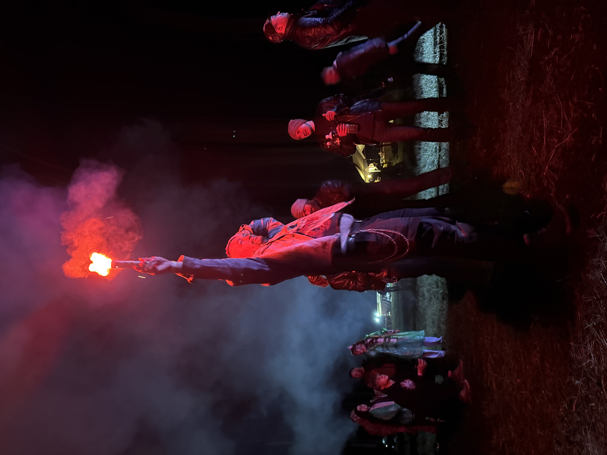 The image displays an outdoor nighttime gathering in Большая Кивара, Russia. A central figure in the foreground, wearing dark clothing and a chain accessory, stands with their right arm extended vertically, holding a bright red handheld flare that emits dense red smoke. The flare's intense light casts a strong red illumination over the immediate surroundings and the individuals present.
Approximately 10-15 people are observable in the midground and background. To the left of the central figure, a person in a dark hoodie is crouching, while another person stands nearby, clad in a translucent green poncho. Further left, a male figure with a beard is visible, illuminated by the red light, amongst other less distinct individuals. To the right of the central figure, a man wearing a dark jacket and a light-colored beanie stands holding a clear plastic bottle. To his right, another man in a dark leather jacket and a dark cap is positioned, looking towards the right side of the frame. A blurred figure is also visible on the far right, suggesting motion.
The ground consists of an unpaved area covered in sparse grass or straw, uniformly colored red by the flare's light. In the distant midground, a white tent-like structure is discernible, emanating a soft yellow internal light. The background features dark silhouettes of trees against the night sky. Red smoke from the flare is distributed throughout the scene, creating a hazy atmosphere, particularly noticeable around the flare and in the upper left quadrant of the image.