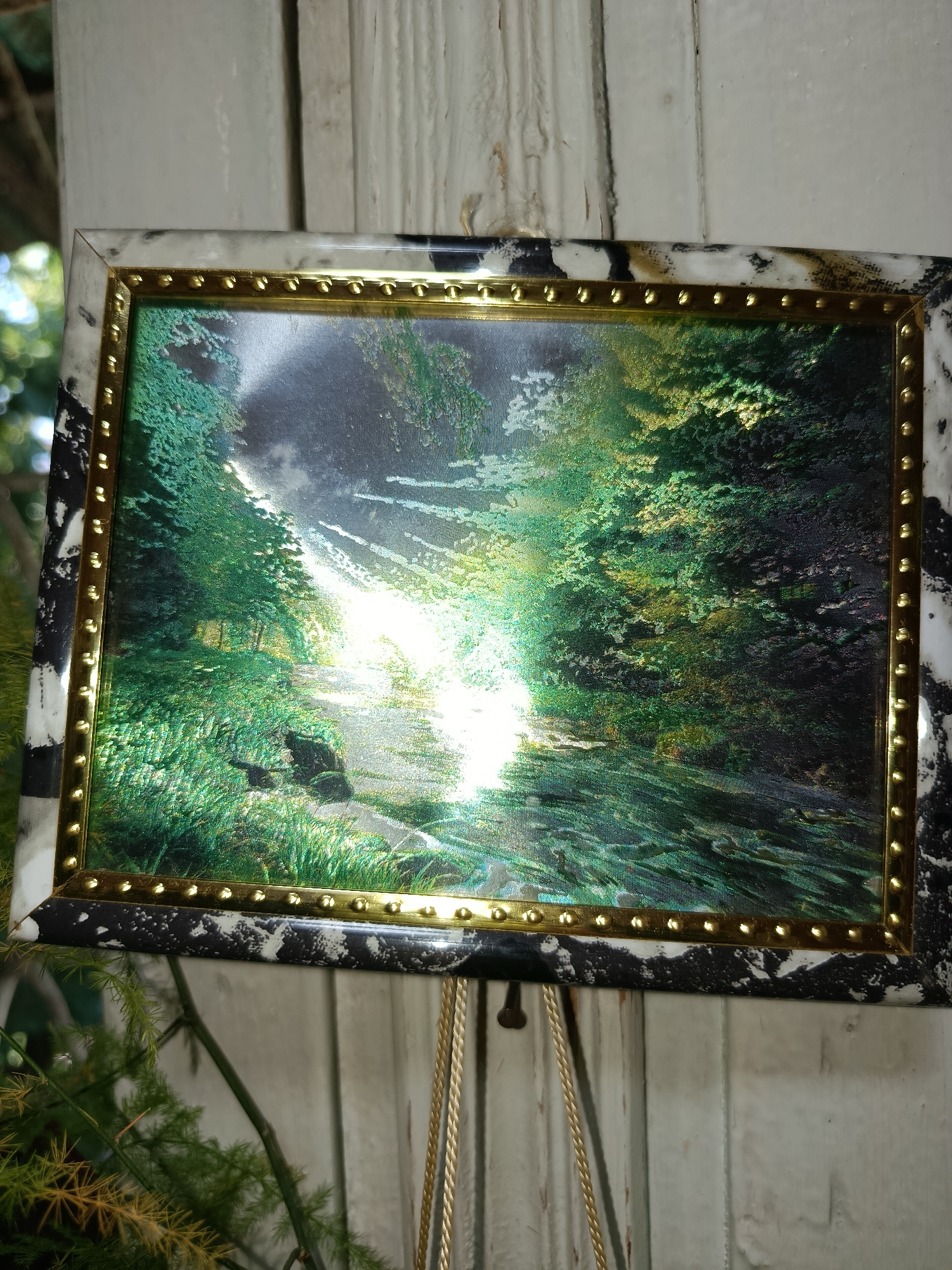 A framed painting of a sunlit forest scene hangs on a wooden surface. The painting depicts a path winding through lush green trees with sunlight streaming through the canopy. The frame has a black and white marbled pattern with a gold beaded inner border. The wooden surface appears to be weathered or painted white, with vertical planks. Green foliage from a plant is visible in the foreground. The overall setting suggests an indoor display, possibly in a home or shop. There is no discernible connection to Tashkent, Uzbekistan, as there are no specific landmarks or cultural elements that identify the location.