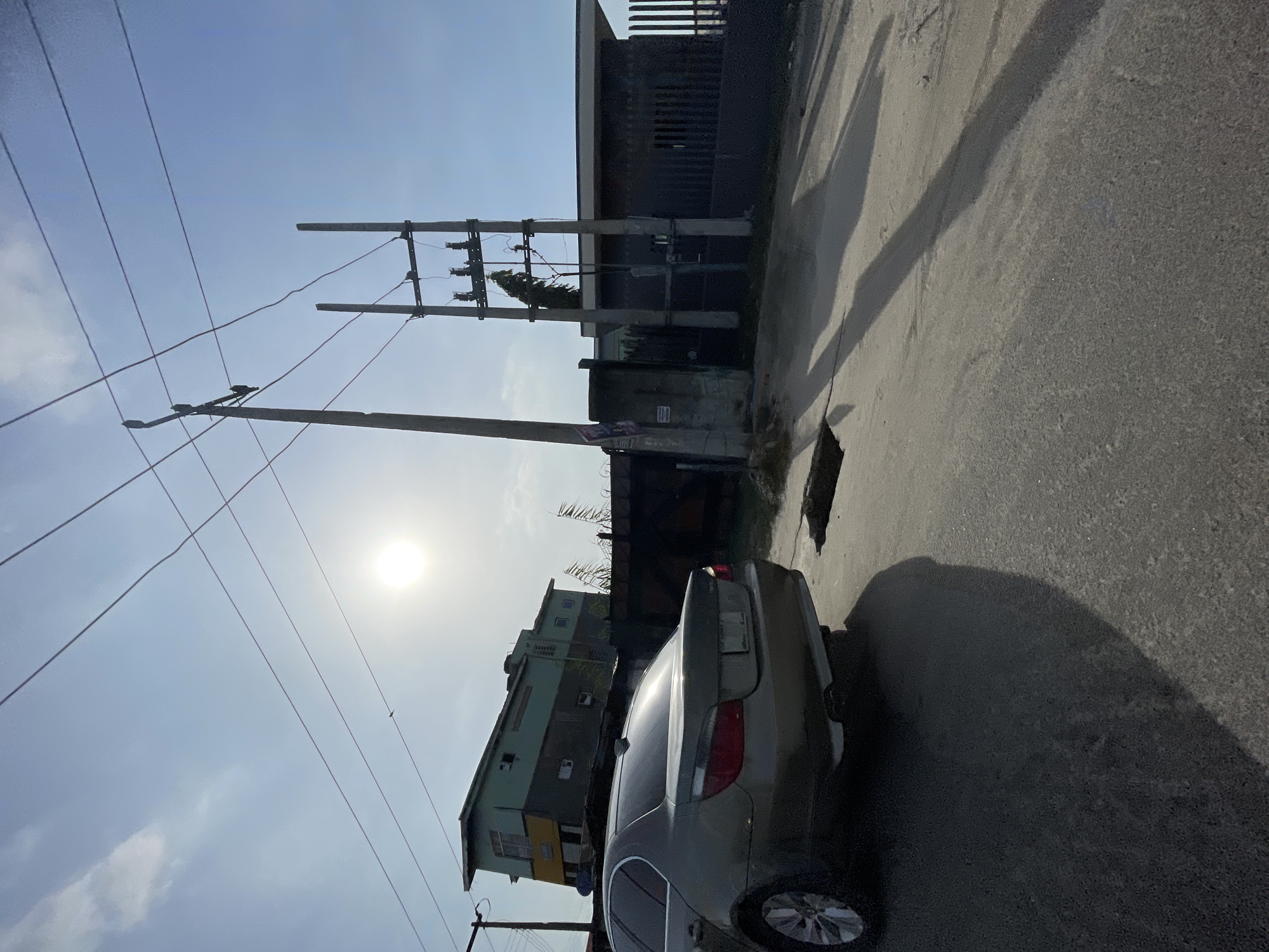 The image depicts a street scene in Ikorodu, Nigeria. A silver sedan is parked on the right side of the asphalt road, next to a visible pothole. Power lines and utility poles are visible on the left, extending upwards towards a bright sky and the sun. In the background, there's a multi-story building with a faded paint job, as well as a taller, darker structure that could be industrial or commercial. The lighting suggests it is daytime. There are no visible people.