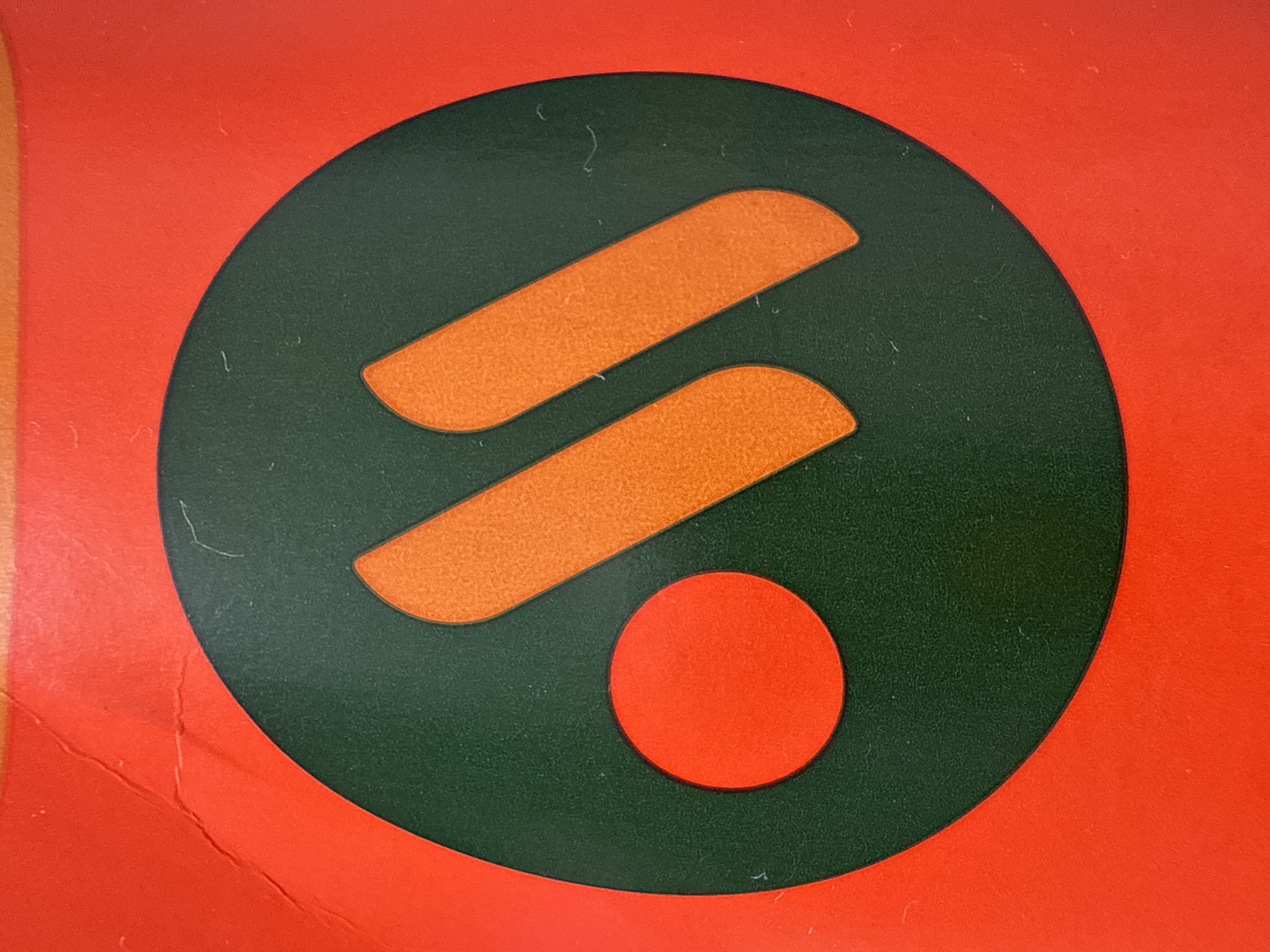 The image shows a close-up of a circular logo with a dark green background. Inside the circle are two orange, elongated shapes that resemble the letter 'M' or two parallel strokes, and a smaller, bright orange circle to the left of the strokes. The logo is set against a vibrant orange background that appears to be some form of textured material, possibly paper or plastic, with visible surface imperfections such as faint lines and scratches. This logo is associated with the Russian fast-food chain "Teremok," known for its blini and other Russian specialties, which has numerous locations across Russia, including in Nizhny Novgorod. The specific location within Nizhny Novgorod cannot be determined from the image alone, as it only depicts the logo.