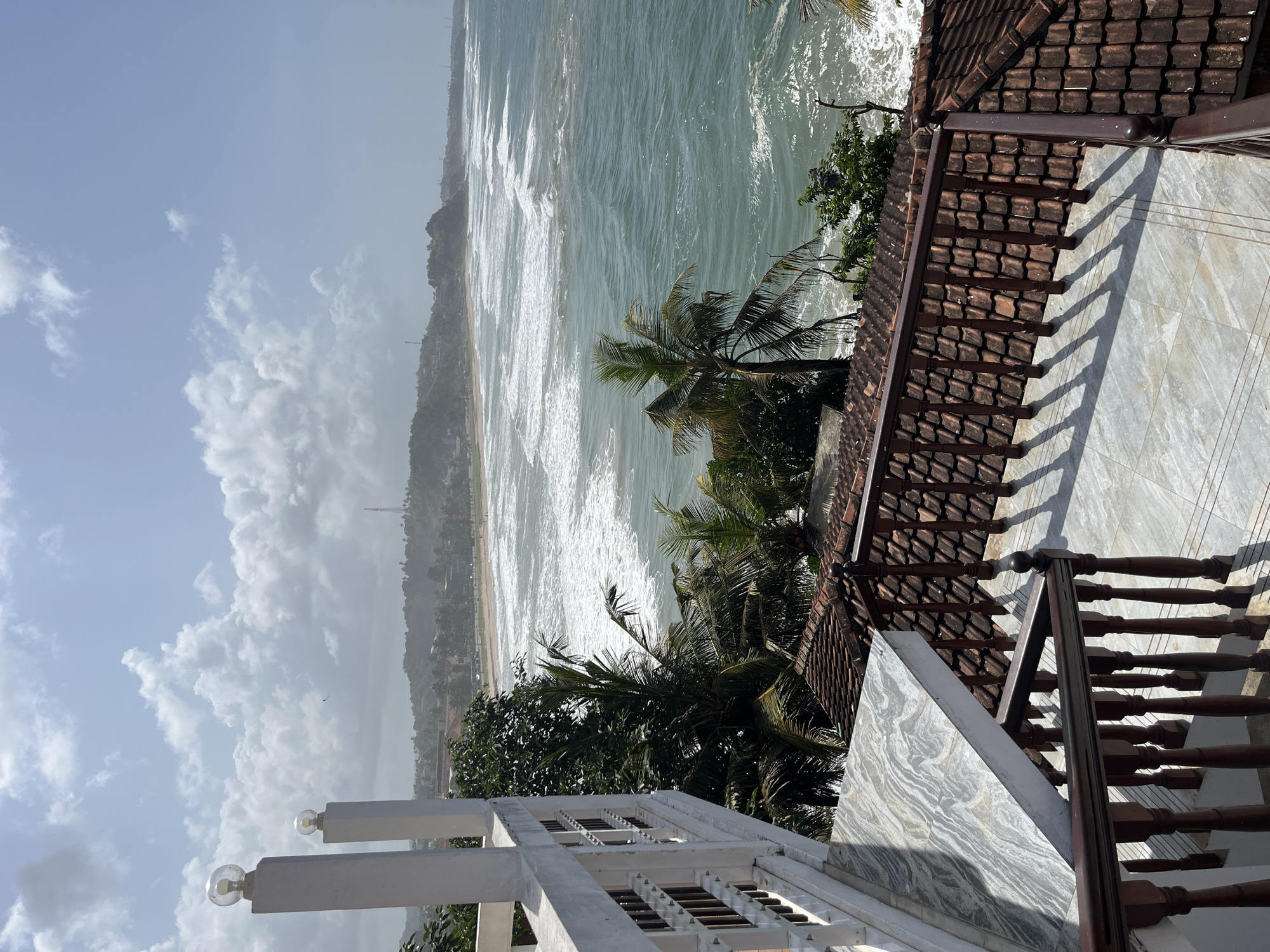 This image depicts a view of the Indian Ocean from an elevated vantage point in Walgama, Sri Lanka. In the foreground, a white balcony railing with vertical dark wooden spindles is visible, casting shadows on a tiled floor. To the right, a tiled roof with reddish-brown tiles slopes downwards. Beyond the immediate structures, lush green palm trees and other vegetation are interspersed. The ocean stretches into the distance, with white-capped waves rolling towards a sandy beach. Along the coastline, a town or village is visible with buildings and more palm trees. The sky is partly cloudy, with white cumulus clouds against a pale blue backdrop. A tall communications tower can be seen on a distant hill. The scene captures a tropical coastal landscape with elements of human habitation.