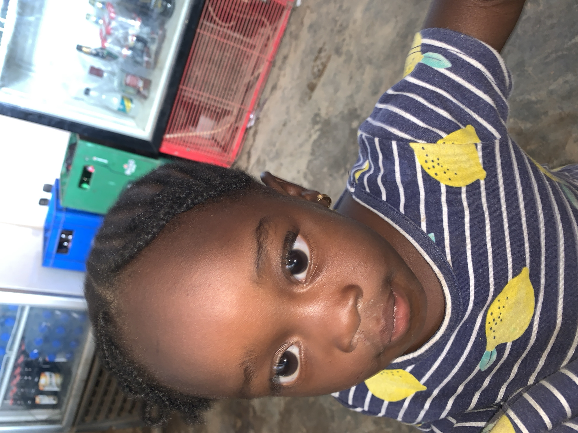 A young girl is looking up at the camera. She has dark skin, brown eyes, and her hair is styled close to her head. She is wearing a navy blue and white striped shirt with yellow lemon designs. In the background, there is a drink cooler with a glass door and various beverages inside. Adjacent to the cooler, there are some crates and containers in shades of blue, green, and red. The scene appears to be indoors, possibly a shop or store. The location is New Stadium, Nigeria.