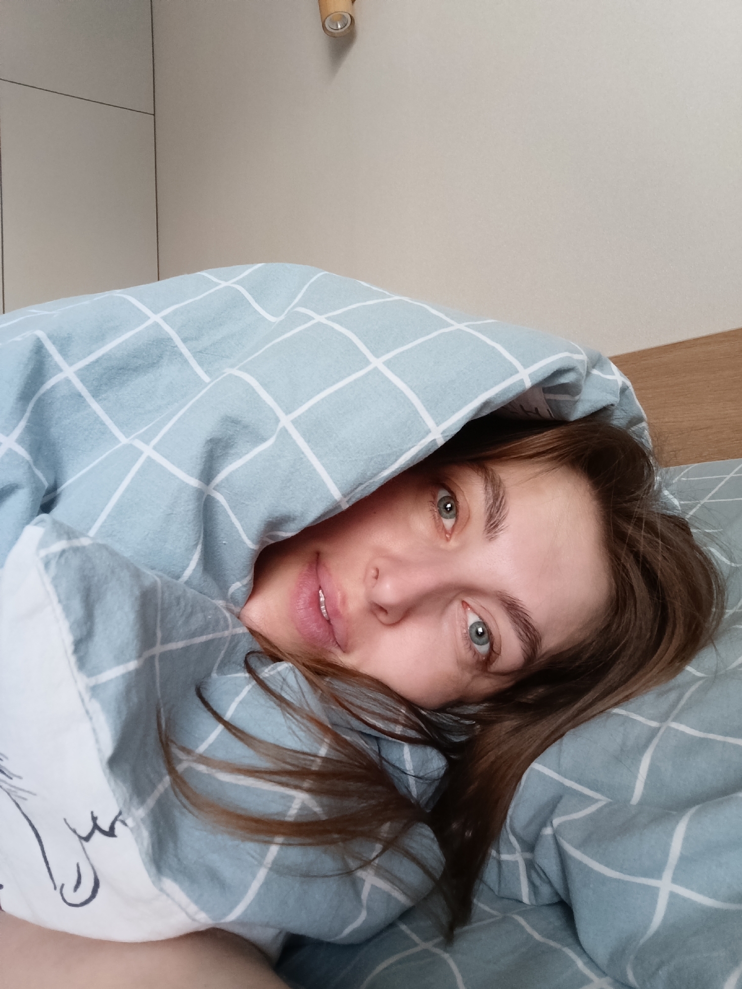 A woman lies in bed under a blue checkered duvet. Her head is slightly tilted to the right, and her eyes are looking directly at the camera. Brown hair is visible around her face, and she appears to be inside a bedroom. There is a wall panel behind the bed and a wooden light fixture in the upper left corner of the image. The city of Chaikovsky, Russia is not directly detectable from the image.