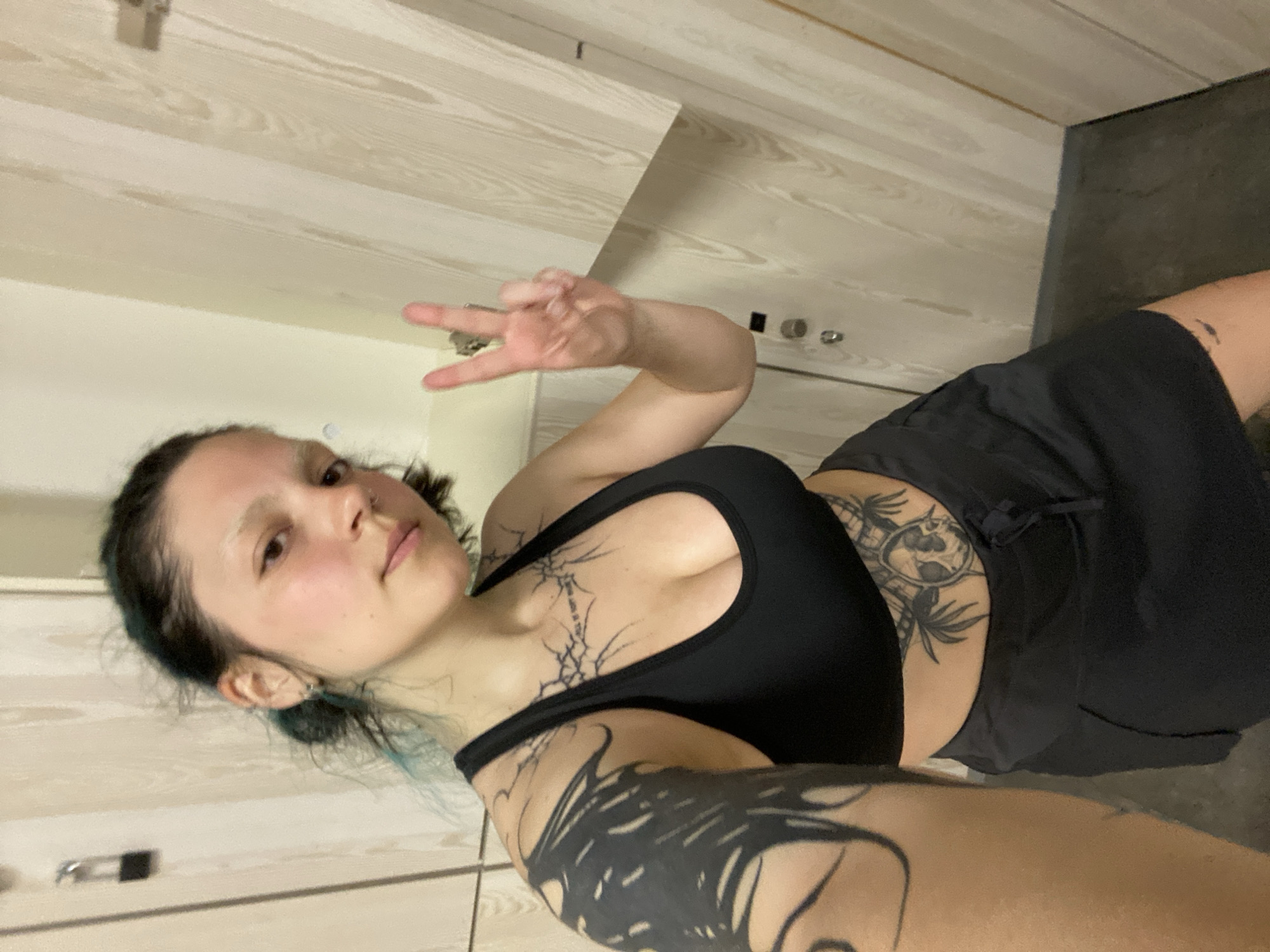 A woman with light skin and dark hair with blue highlights is posing for a selfie in what appears to be a bathroom. She is wearing a black sports bra and dark shorts. Prominent tattoos are visible on her neck, arms, and torso. She is making a peace sign with her left hand. The background includes light-colored wood-paneled walls and a gray floor. It's unlikely to determine if the location is in Gijón/Xixón, Spain, without further information.