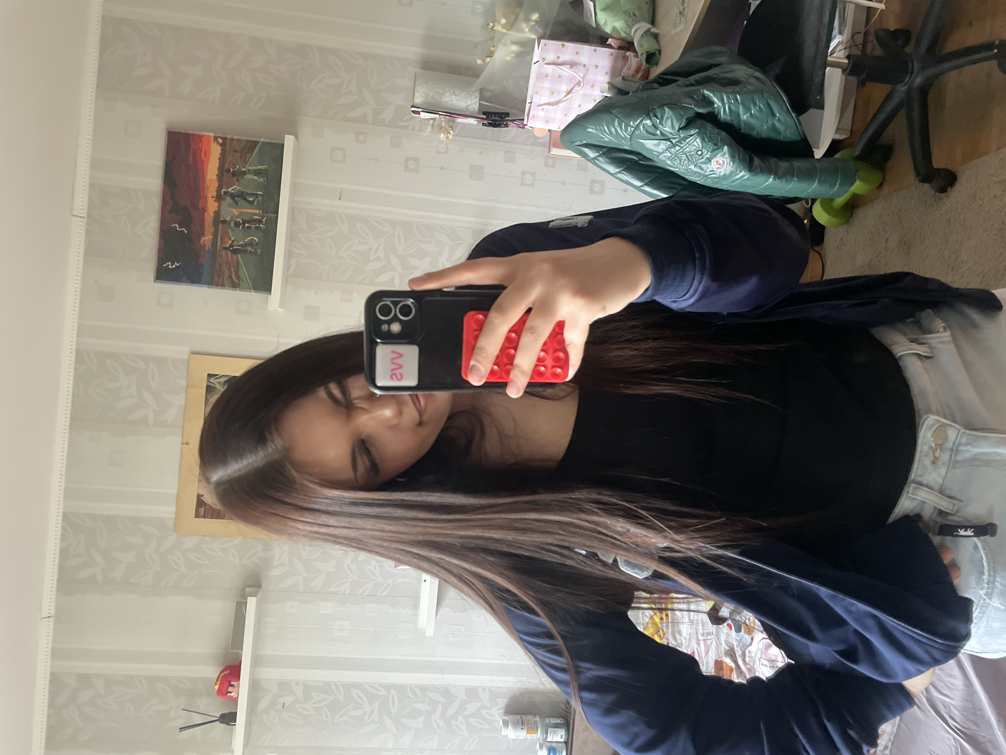 A young woman captures a mirror selfie in an indoor setting, likely a room in Vsevolozhsk, Russia. She has long, dark brown hair and is dressed in a black top, a dark blue zipped jacket, and light-wash jeans. Her head is tilted slightly, and her facial expression appears neutral or mildly pleasant.
She holds a black smartphone with a dual-camera setup, a red pop-it style grip on the back, and a white sticker displaying the text "2VV" (which would read "VV2" if seen directly).
The background, reflected in the mirror, reveals light-colored wallpaper adorned with a subtle, repeating leaf or floral pattern. Various objects are visible: a framed painting depicting horses and birds against a dramatic sunset or sunrise sky, a collection of gifts or wrapped items including a pink patterned bag, and a dark green quilted Moncler puffer jacket with its distinctive logo patch. Below these items, a black office chair and a green dumbbell are on a carpeted floor. On a lower surface or shelf, several white plastic bottles, possibly containing medicine or supplements, can be seen. Part of a bed with light-colored bedding is also visible in the lower right corner of the image. The scene suggests a personal living space.