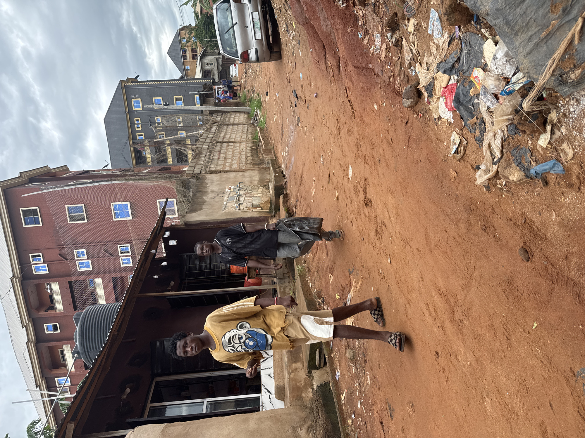 The image shows a dirt road in Nkpor, Nigeria. Two young men are walking on the road. The man in the foreground wears a yellow t-shirt with a cartoon graphic and light-colored shorts. The man in the background wears a black t-shirt and denim jeans, and carries a black plastic bag. A building sits to the left of the road, and trash is piled along the right side of the road. A silver car is parked further up the road, and several buildings are visible in the background. The sky is overcast.