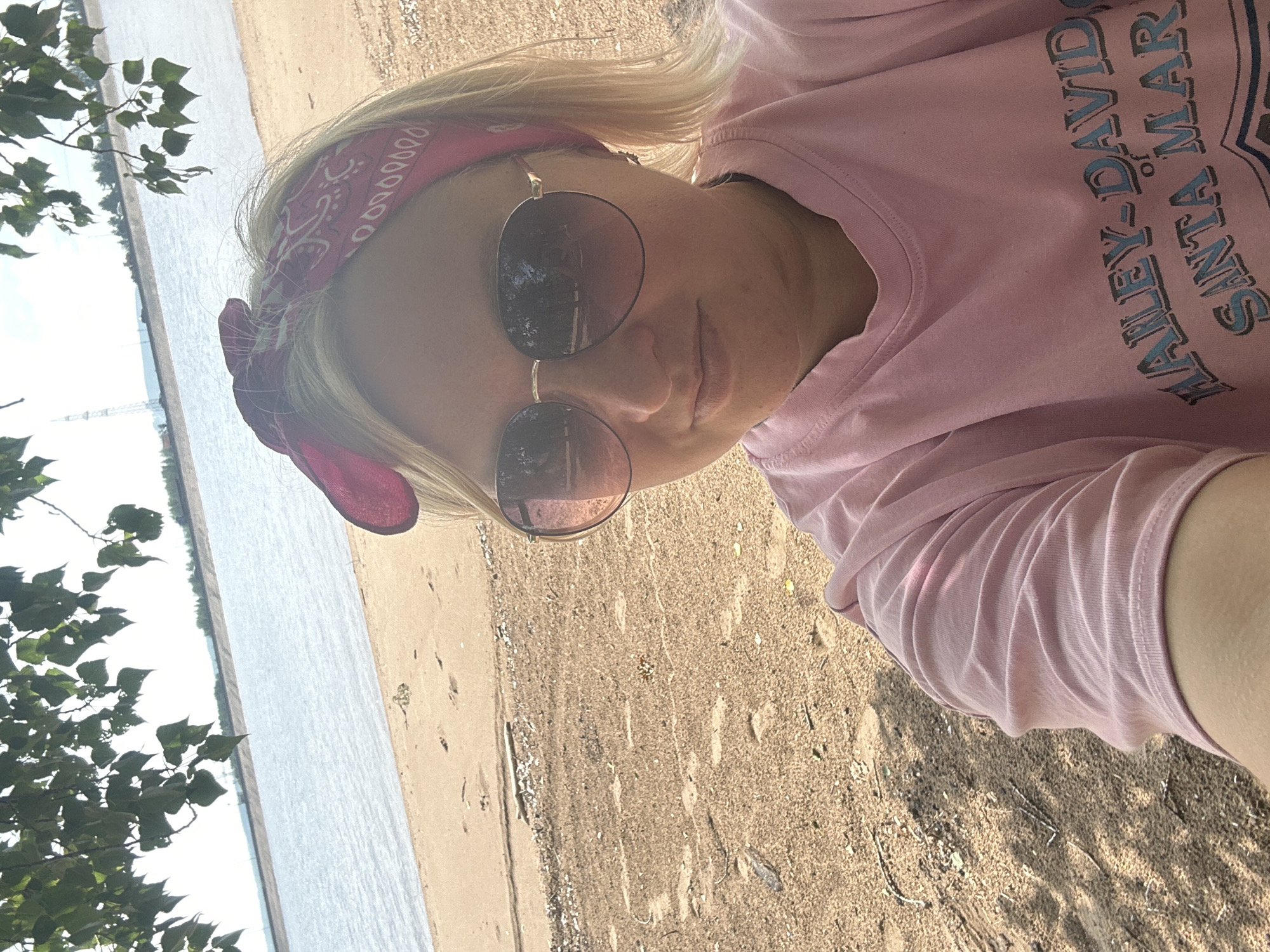 A blonde woman wearing a pink bandana and sunglasses takes a selfie on a sandy beach near the water. She is wearing a pink t-shirt with blue lettering that reads "HAILEY-DAVID of SANTA MAR". The beach is strewn with debris and has footprints. In the background, a body of water is visible, with a distant shoreline and a clear sky. The image is shot from a slightly low angle, with tree branches and leaves in the upper left corner.