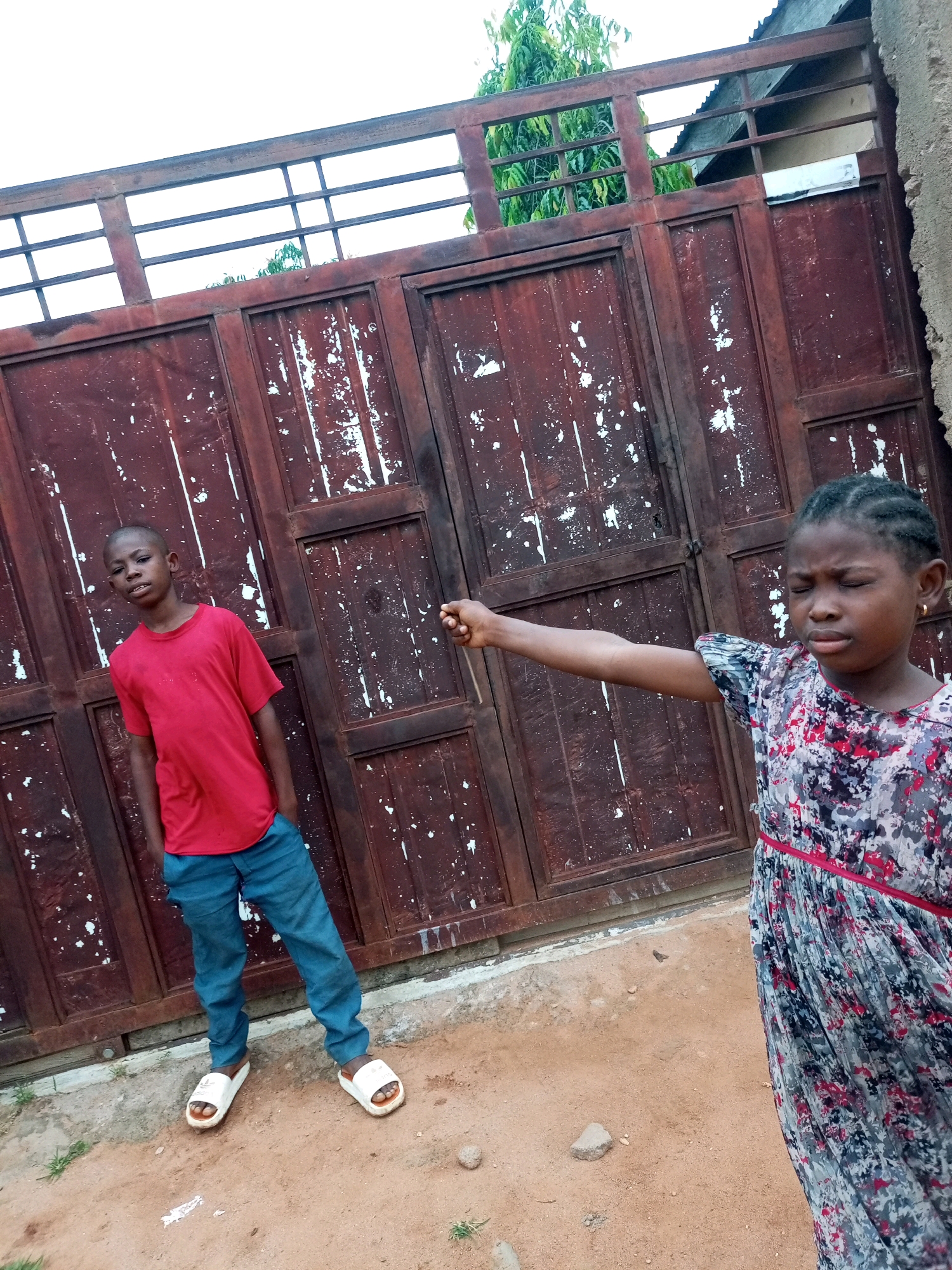 A full-body outdoor shot shows a young Black boy and a young Black girl standing in front of a large, weathered, dark red metal gate with horizontal bars at the top. The boy, on the left, is wearing a bright red t-shirt and blue jeans, with his hands in his pockets and white slip-on sandals. He is looking towards the camera with a slight smile. The girl, on the right, is wearing a gray, pink, and black floral print dress with a red belt. Her right arm is extended outward, and she appears to be gesturing or pointing. Her eyes are closed, and her mouth is slightly open. The ground in front of the gate is a mixture of dirt and some sparse green grass. The gate shows signs of age and wear, with areas of peeling paint revealing a lighter color underneath. The background above the gate includes some green foliage from trees and a glimpse of a building's roof. The lighting suggests it is daytime, possibly overcast. The scene is in Tempure, Nigeria.