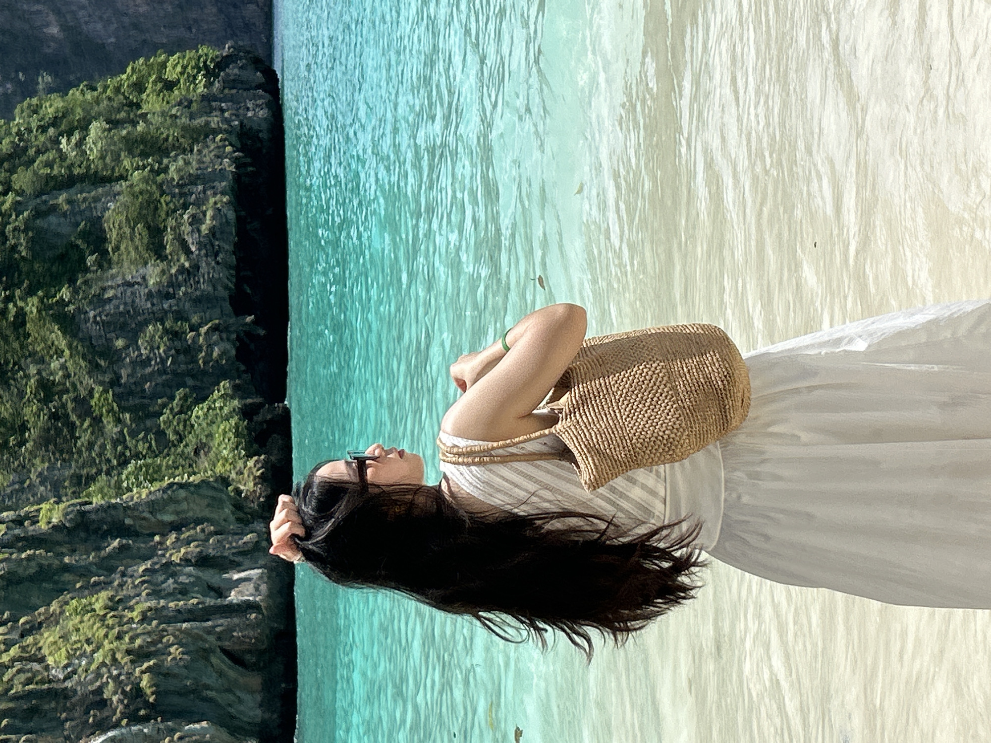 An outdoor, medium shot captures a woman standing on a sandy beach at the edge of clear turquoise water, with a lush, rocky cliff face in the background. The woman, facing away from the camera and looking out at the sea, has long dark hair, sunglasses, and wears a white dress and a straw shoulder bag. She is touching her hair with her right hand. The water is calm with gentle ripples, and a few small objects are visible in the shallow water. The cliff face is covered in dense green vegetation. The scene suggests a tropical beach destination, likely in the Ao Nang area of Thailand, known for its limestone karsts and clear waters.