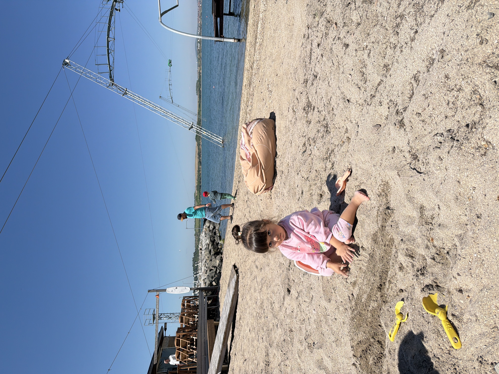 A young girl in pink pajamas sits on a sandy beach, playing with yellow plastic toy shovels. To her left is a brown bag or rolled-up blanket. In the background, two people stand near a rocky shore by the water, under a clear blue sky. A wakeboarding cable lift structure is visible in the background, along with other structures that indicate this is likely a commercial beach or water sports area. This image was likely taken on the beach near Burgas, Bulgaria, based on the architectural style of the waterfront structures.