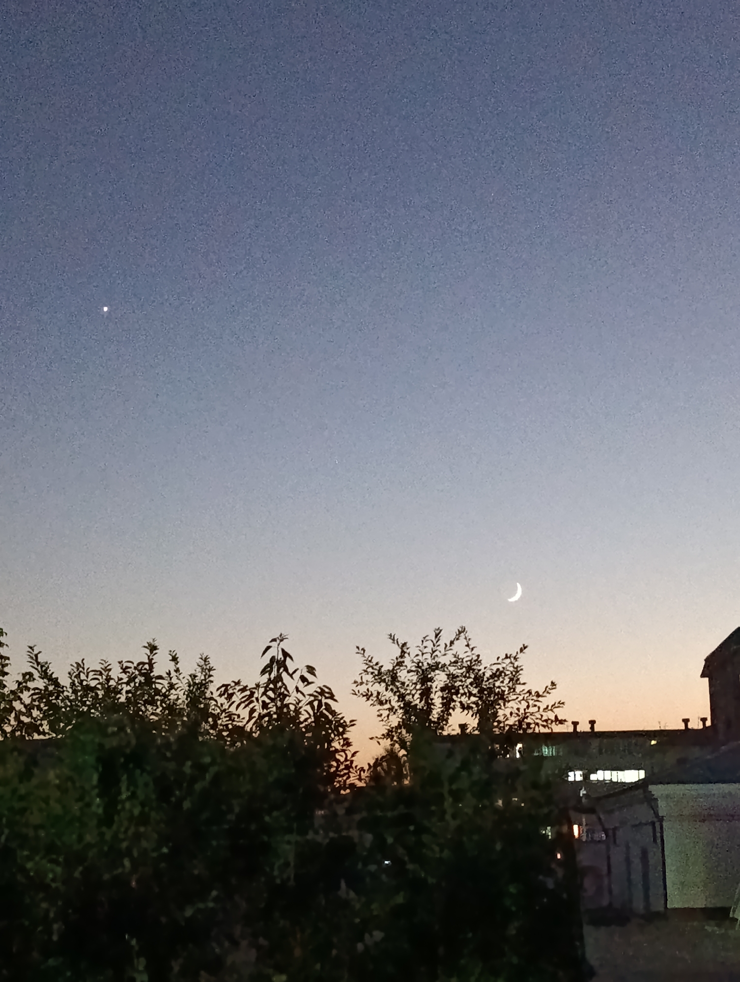 The image depicts a twilight sky over Fergana City, Uzbekistan, with a crescent moon visible on the right and a bright star or planet on the left. Silhouetted trees and buildings form the foreground. The sky transitions from a deep blue at the top to a lighter, warmer hue near the horizon. The overall scene is calm and serene.