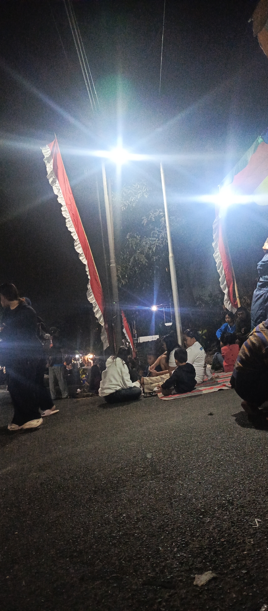 In the city of Sumbersekar, Indonesia, at night, several people are gathered on an asphalt road. Bright lights, possibly from streetlamps or temporary lighting, illuminate the scene, causing lens flare. Red and white striped flags are prominently displayed, attached to poles. Some individuals are sitting on the ground, while others are standing. The atmosphere appears to be a community gathering or event taking place outdoors after dark.