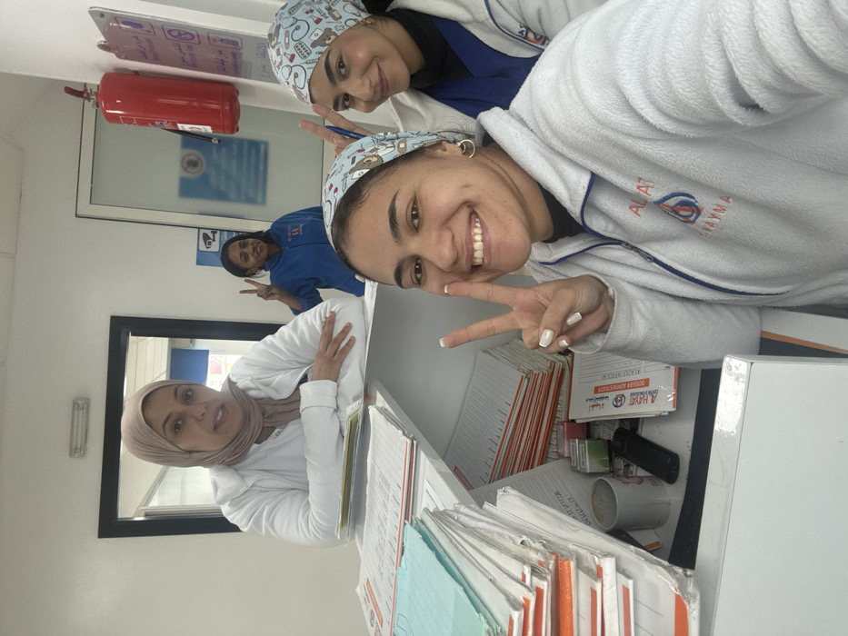 Four adult females are depicted in a well-lit indoor reception or administrative office of a medical facility in Meknes, Morocco.
In the foreground, two women pose smiling behind a white counter. The woman on the left, with dark hair, wears a white fleece jacket bearing the "ALAT ANAYMA" logo with a blood drop symbol, and a light blue patterned scrub cap. She holds her right hand up in a peace sign. The woman on the right, also with dark hair, wears a similar patterned scrub cap and makes a peace sign with her left hand while holding a pen.
Behind the counter, a third woman, wearing a beige hijab and a white lab coat, rests her forearms on the counter, smiling toward the camera. Further in the background, a fourth woman with darker skin is visible through a doorway opening; she wears a blue zip-up jacket with "AL-GHAYAT" visible on the chest and also makes a peace sign.
The white counter is occupied by numerous stacks of documents, some with orange borders, others light blue, many bearing Arabic text and "DOSSIER MEDICAL" (medical file). A white mug, small office supply containers, and a black remote control are also present on the counter. The background features white walls, a mounted red fire extinguisher, and multiple wall-mounted signs with Arabic text and symbols, including a security camera icon and medical equipment illustrations. A fluorescent light fixture is visible on the ceiling. All individuals are posing, indicating a group photograph.