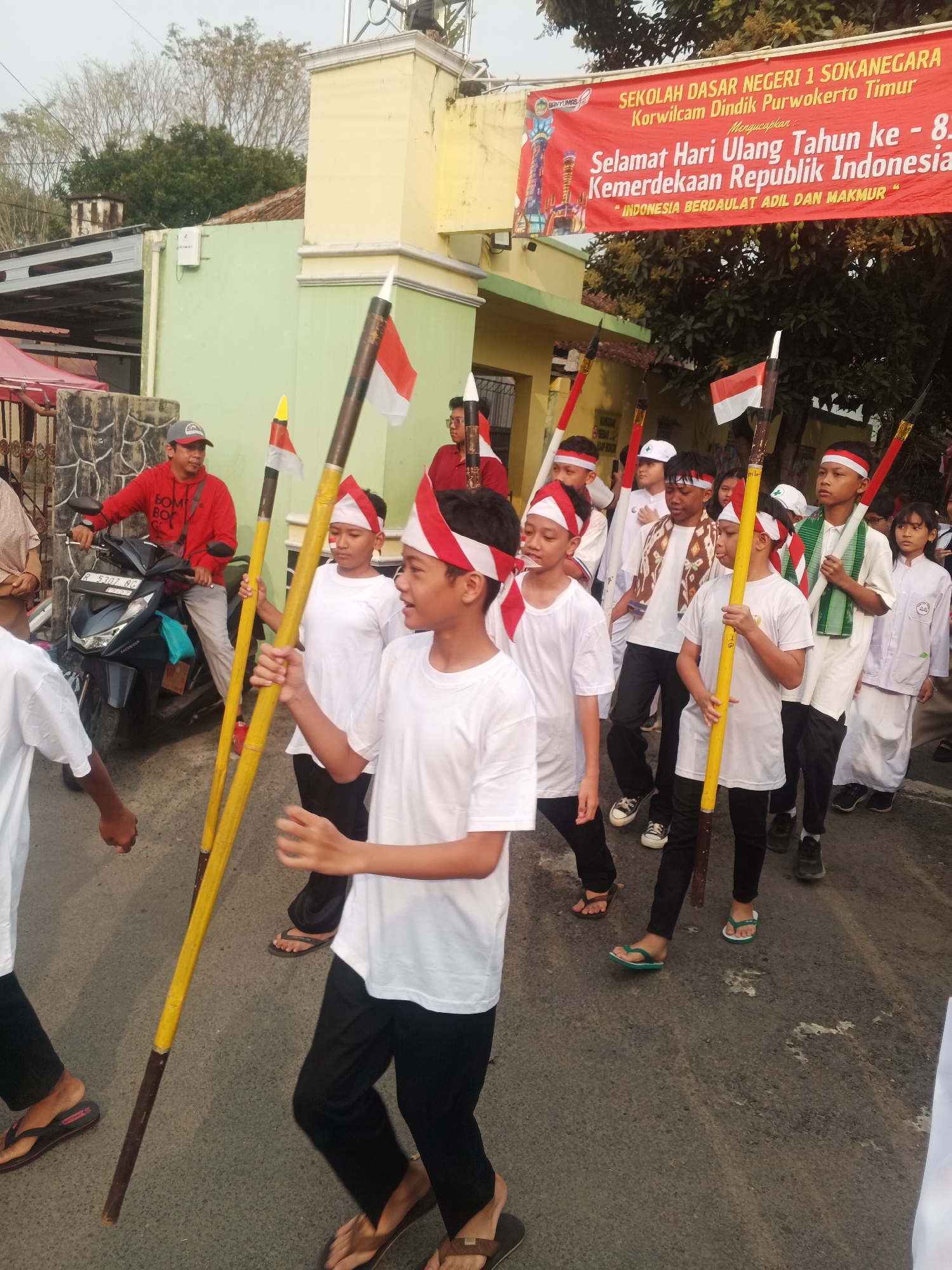 In Sokanegara, Indonesia, a group of schoolchildren are participating in a parade to celebrate Indonesia's Independence Day. The children, dressed in white shirts and headbands, are carrying poles adorned with Indonesian flags. A banner overhead, visible in the upper part of the image, reads "SEKOLAH DASAR NEGERI 1 SOKANEGARA" and congratulates the audience on "Selamat Hari Ulang Tahun ke - 8... Kemerdekaan Republik Indonesia." The parade appears to be taking place on a street with buildings lining one side and trees in the background. A man on a motorcycle is also present, observing the procession.