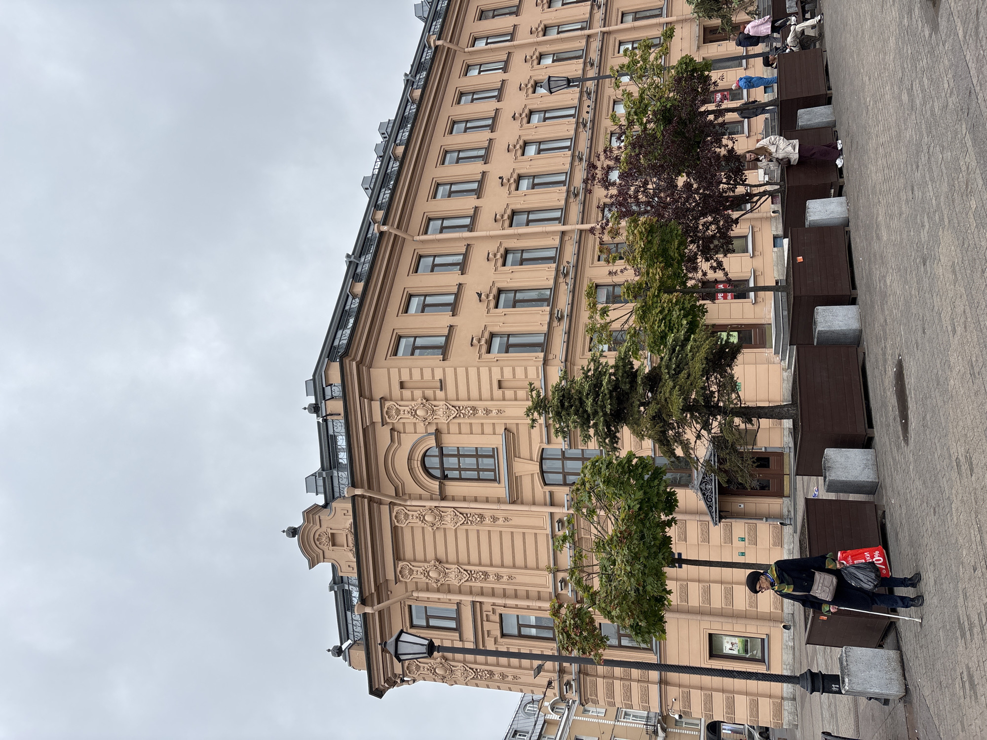 A slightly angled, full outdoor shot depicts a tall, multi-story, light beige building in Saint Petersburg, Russia. The sky is overcast and gray. The building is adorned with decorative architectural details around its numerous windows and at its top. In the foreground, a sidewalk area is visible, marked by dark brown rectangular planters interspersed with square concrete blocks. Trees are planted within these containers, adding greenery to the scene.
A person is walking on the sidewalk. The individual is dressed in dark clothing and uses a white cane for guidance, suggesting they are visually impaired. They also carry a red shopping bag. Several individuals are seated on the benches nearby. A lamppost stands prominently in the foreground.