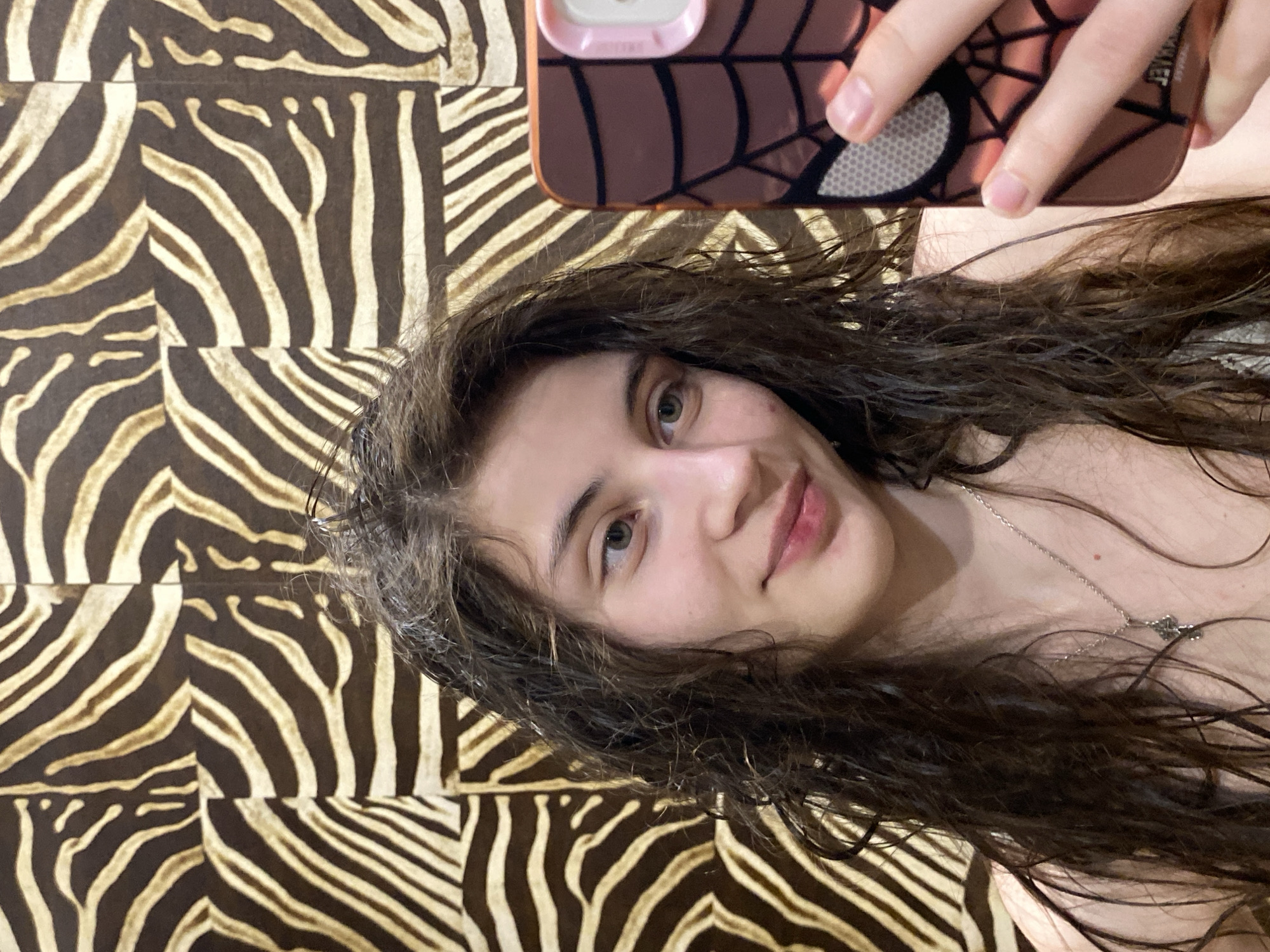 A young woman takes a selfie from an overhead perspective in Votkinsk, Russia. She is lying down, looking up at the camera with a calm expression and a gentle, subtle smile. She has long, dark, wavy hair that frames her face and partially covers her chest, where a delicate silver-colored chain necklace with a small pendant is visible. Her eyes are light-colored, possibly green or hazel. She holds a smartphone above her with her right hand; the phone case is brown/bronze with a distinct spiderweb pattern, a pink edge, and a visible circular detail. The background behind her is a bold dark brown and light beige zebra-print pattern, likely a blanket or bedding. The scene is indoors, under what appears to be ambient lighting. No specific time of day or weather conditions are discernible, and no significant readable text is visible.