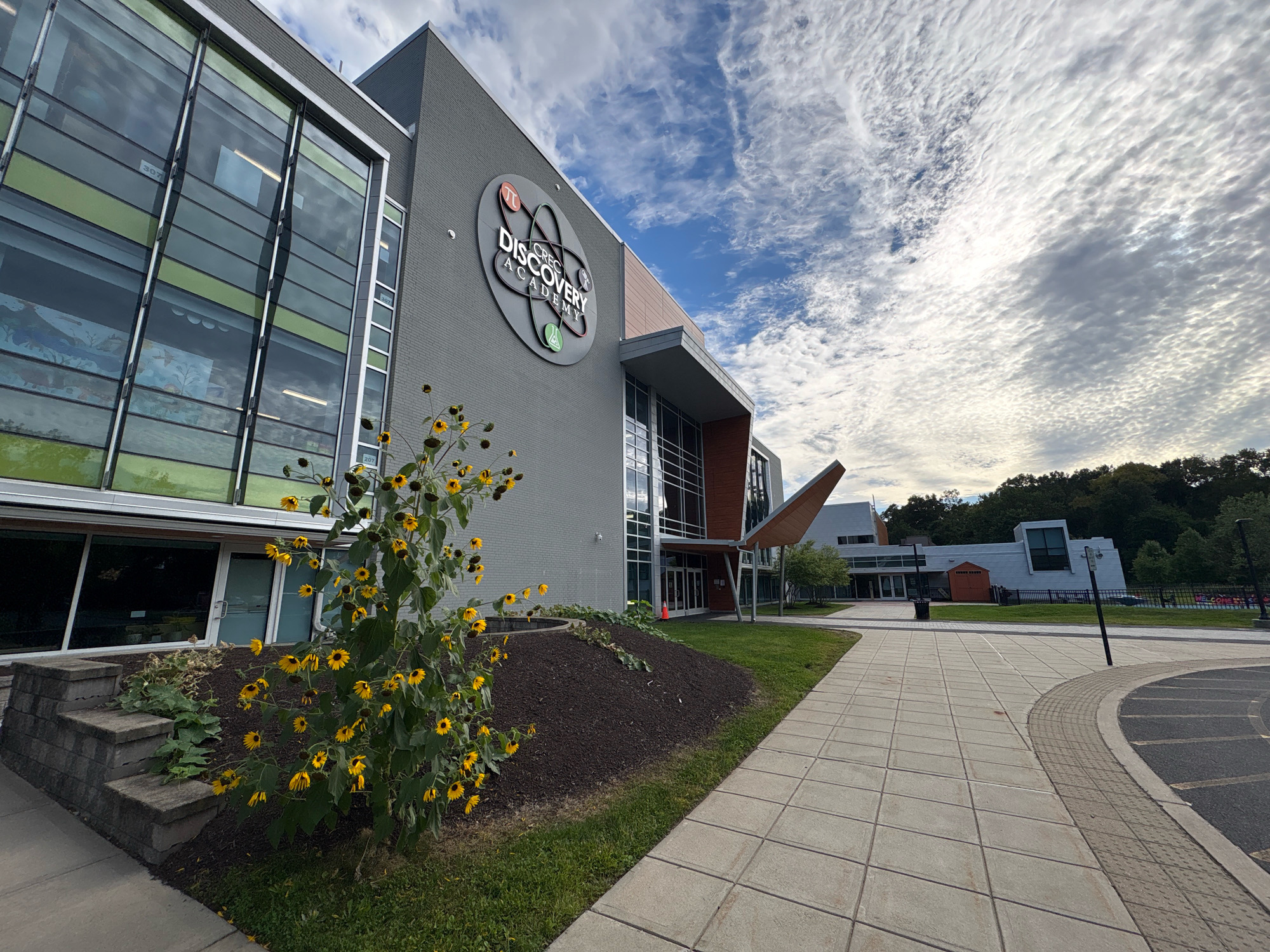 This image shows the CREC Discovery Academy in Wethersfield, Connecticut, United States of America. The building features a modern architectural style with a grey brick facade and large glass windows. A circular logo with the words "CREC DISCOVERY ACADEMY" and scientific symbols is prominently displayed on the building's exterior. In the foreground, a bush with yellow flowers is visible, along with a paved walkway leading towards the entrance of the building. The sky above is partly cloudy with scattered white clouds against a blue background. Trees and other buildings are visible in the distance. The scene appears to be captured on a bright, overcast day. There are no people visible in the image.