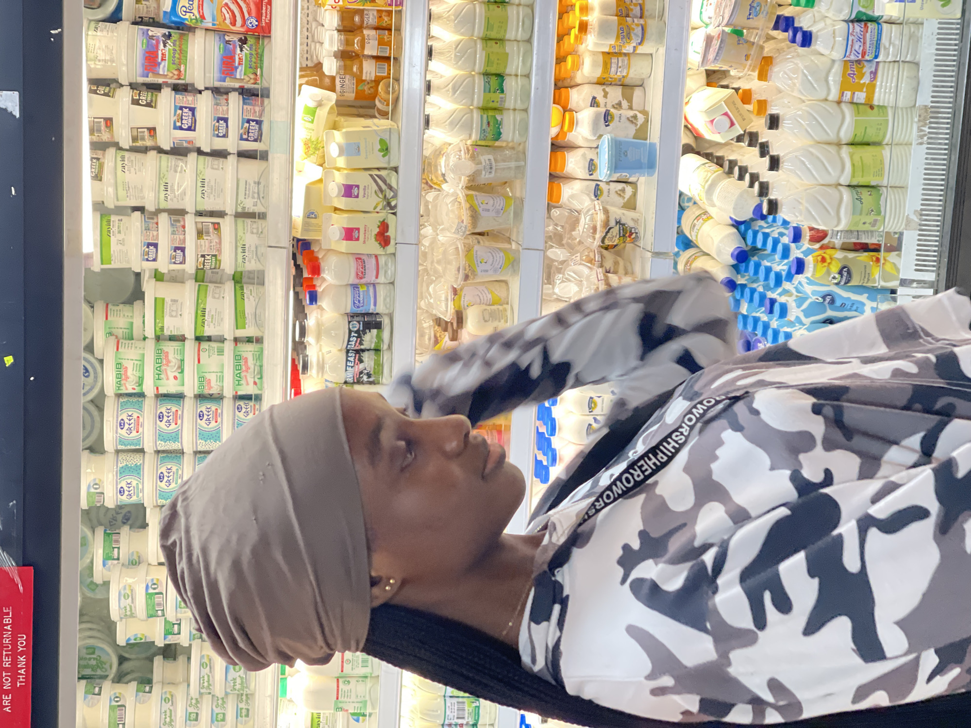 The image shows a young Black woman in a grocery store, likely in Guzape, Nigeria, based on the provided context. She is wearing a headscarf and a camouflage-patterned jacket with the words "WORSHIPHEROWOR" visible on a white strap across the shoulder. The woman is looking to her right, with a neutral expression.
Behind her, the scene is a brightly lit refrigerator aisle filled with various dairy products. Numerous containers of yogurt, labeled "GREEK" and "HABIB," are stacked neatly. There are also several bottles of milk, some with blue caps and labels, and other assorted beverage containers. Some of the bottles appear to be from brands like "Amira" and "Amul." The overall impression is one of a well-stocked grocery store refrigeration section. A red sign with white text that reads "ARE NOT RETURNABLE THANK YOU" is visible on the left side of the image. The lighting suggests it is daytime inside the store.
