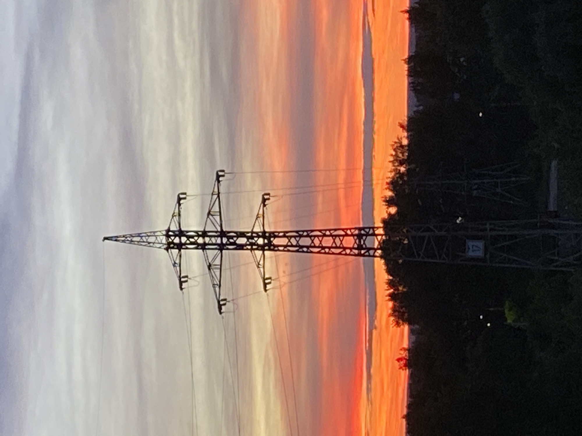A silhouette of a high-voltage electricity pylon stands against a vibrant sunset sky. The sky transitions from a pale gray at the top to fiery oranges and reds near the horizon, with distinct cloud formations visible. The pylon, made of dark metal latticework, is topped with insulators and wires that stretch out of frame. Below the pylon, a dense line of dark green trees forms the horizon, obscuring the ground. The overall scene depicts a somewhat industrial landscape meeting natural beauty at dusk. The specific location in Novokuybyshevsk, Russia, is an area with power infrastructure amidst natural surroundings.