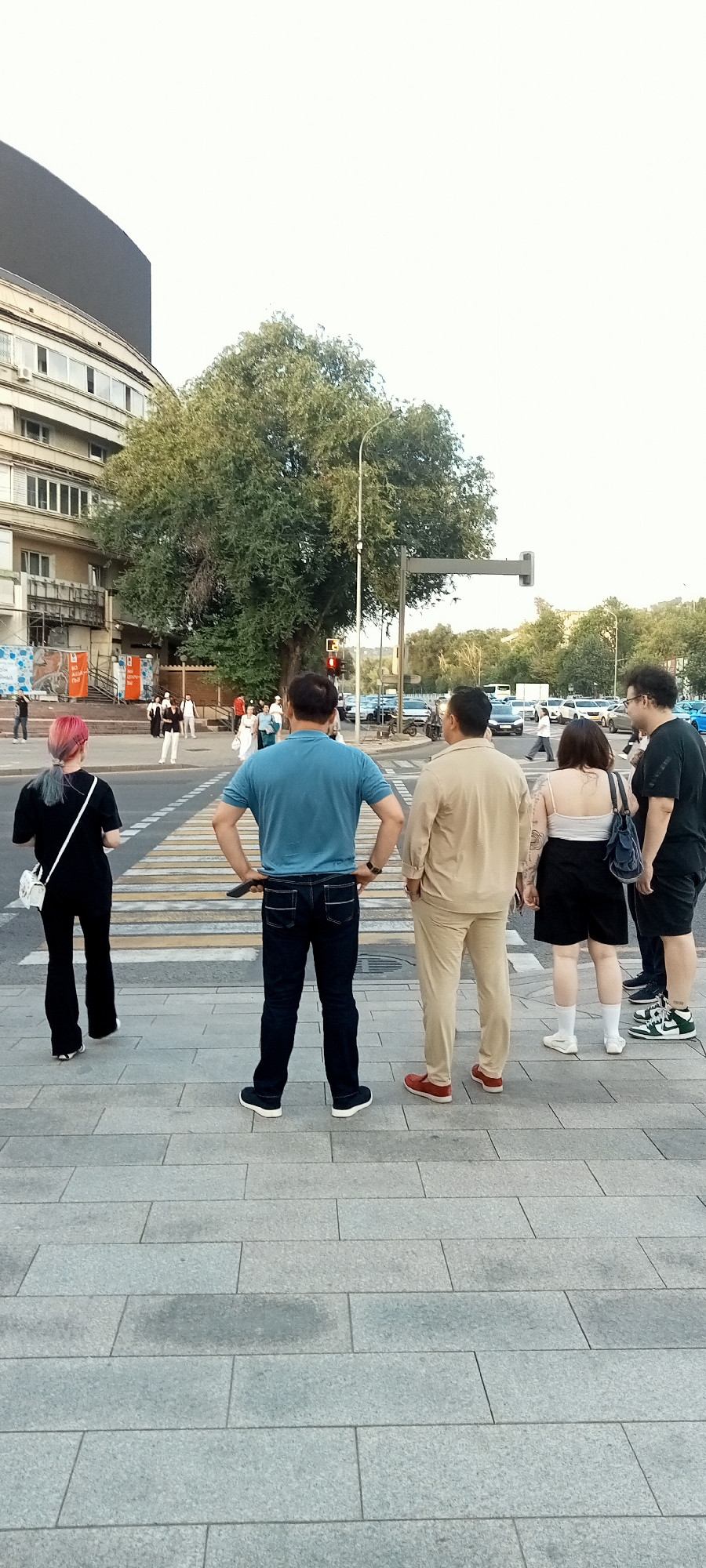 The image shows a street scene in Almaty, Kazakhstan, with a pedestrian crossing in the foreground. Several people are standing on the sidewalk, facing away from the camera. In the background, there are buildings, trees, and traffic. The weather appears to be overcast.
Specifically, the image captures a group of individuals waiting at a crosswalk, suggesting they are about to cross the street. The architectural style of the buildings visible indicates an urban environment, and the presence of trees suggests a well-developed city. The pedestrian crossing itself, marked with white stripes, is a common urban feature. Traffic lights and vehicles are visible in the background, confirming the location as a public street with vehicular traffic. The overall atmosphere is one of everyday urban life.
