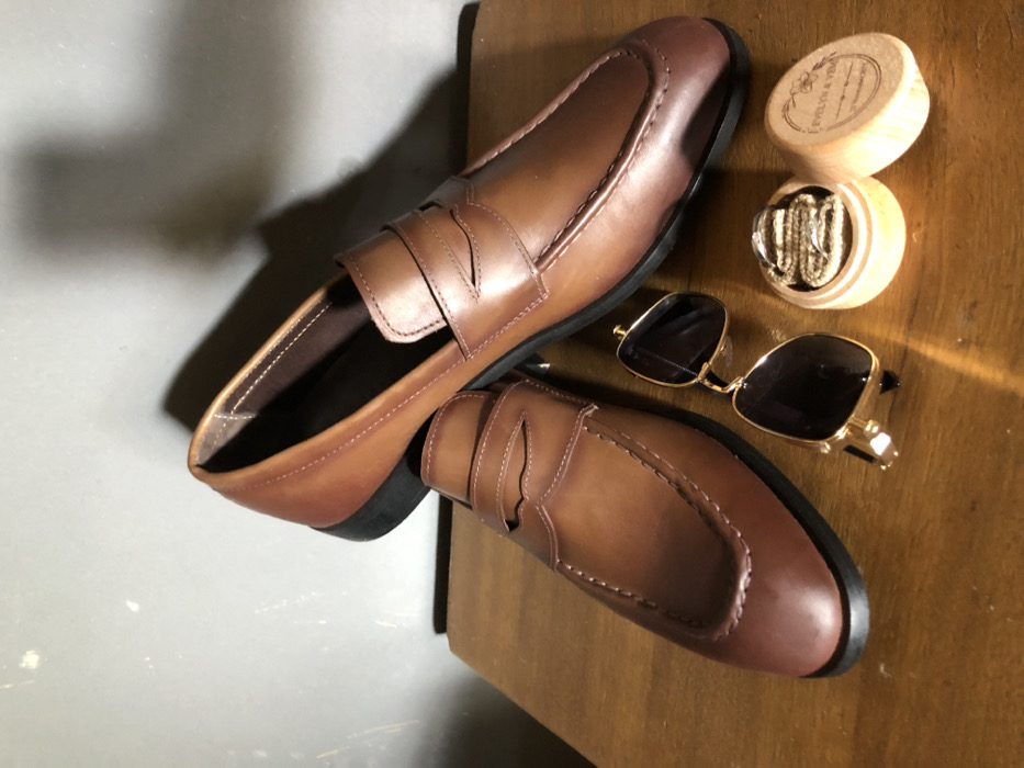 A pair of brown leather loafers rest on a wooden surface, one partially overlapping the other. Adjacent to the shoes are a pair of sunglasses with gold frames and dark lenses. Next to the sunglasses is a small, round wooden box with an open lid, revealing a string or ribbon inside. The scene appears to be set indoors with a plain gray surface visible in the background.